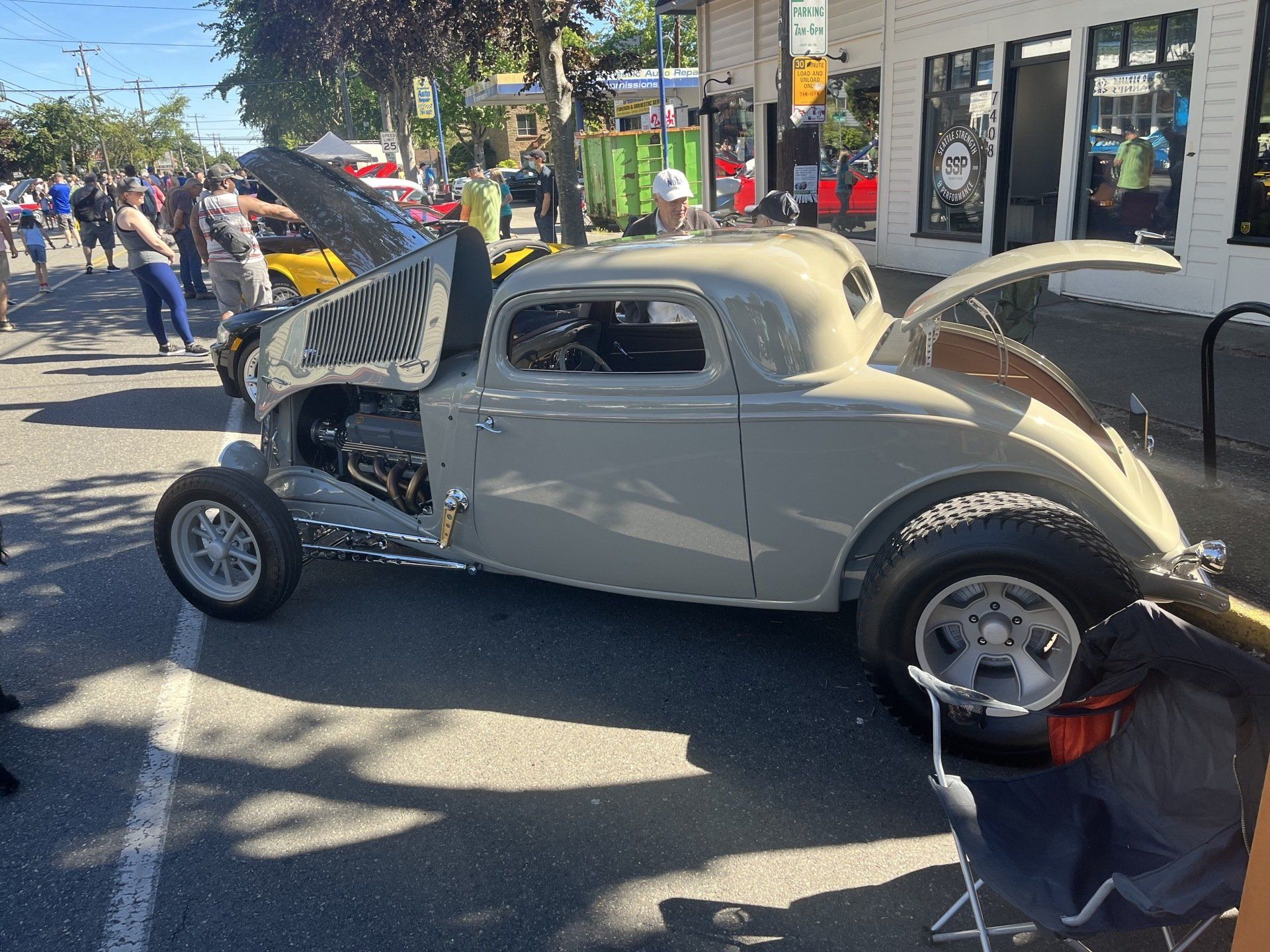 Classic cream-colored hot rod with its hood open at an outdoor car show, people visible nearby.