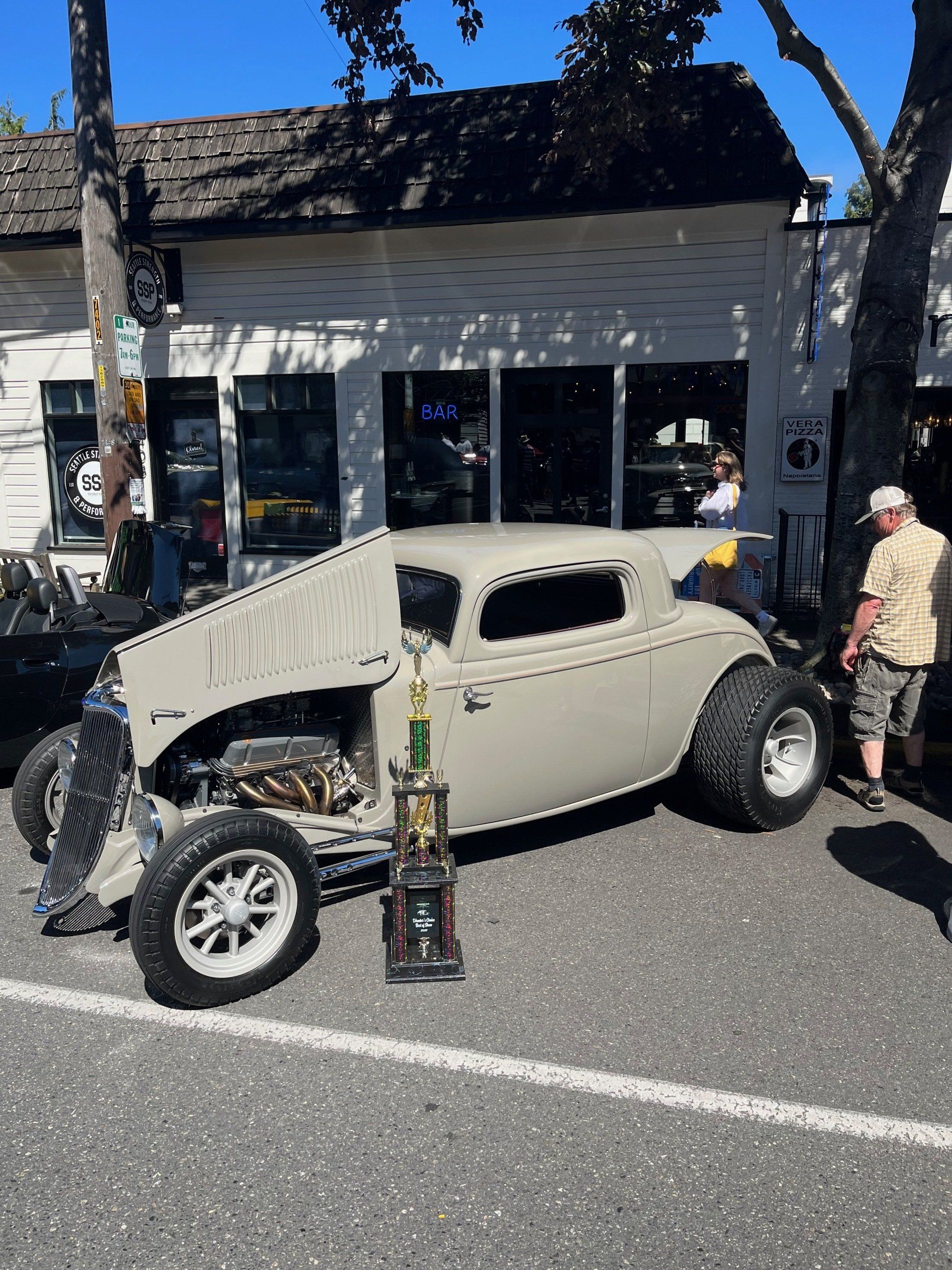 Vintage beige hot rod on a street, with a small trophy in front and people standing nearby.