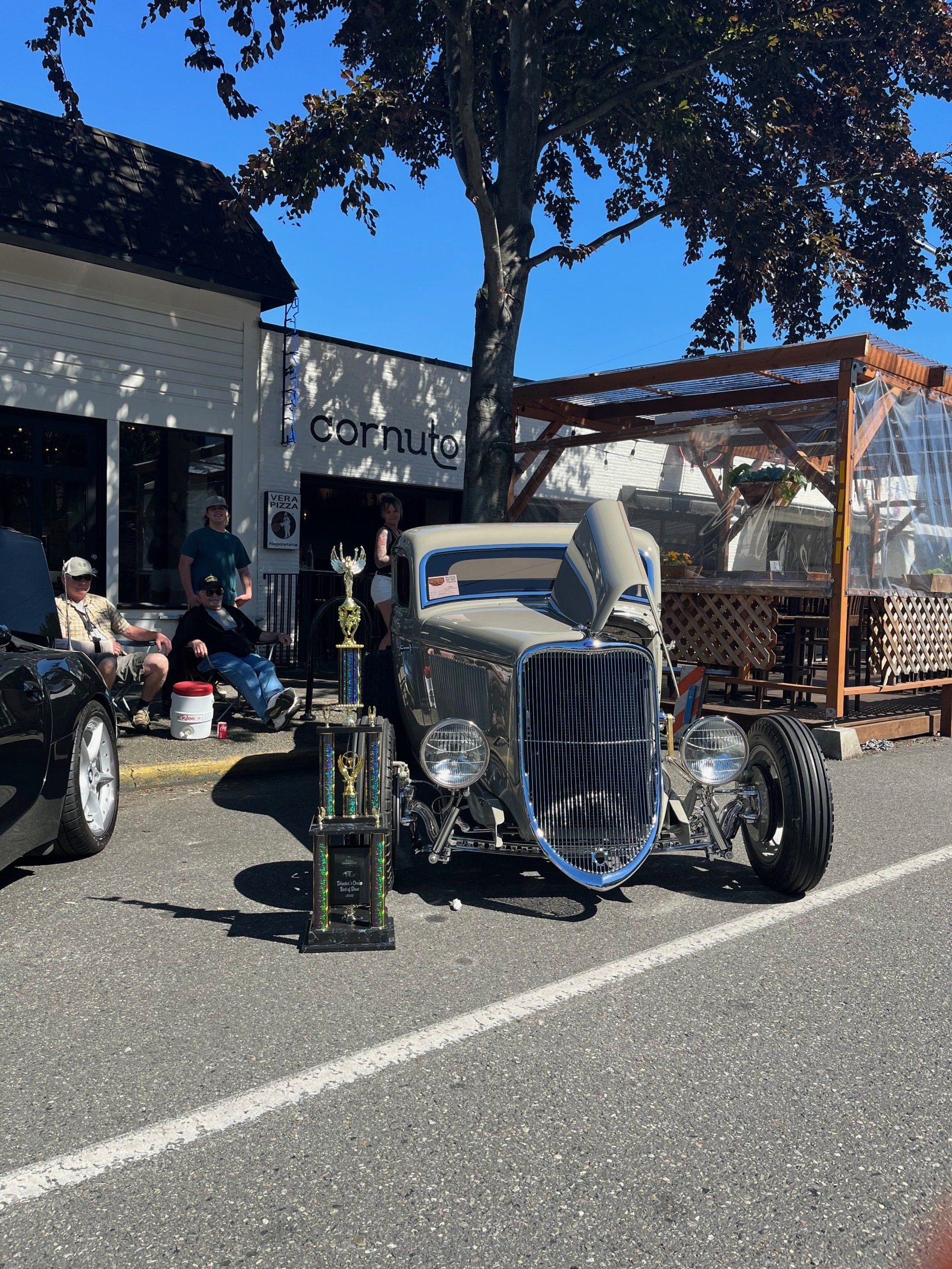 Custom hot rod car parked outside a shop on a sunny day, with trophies displayed nearby.