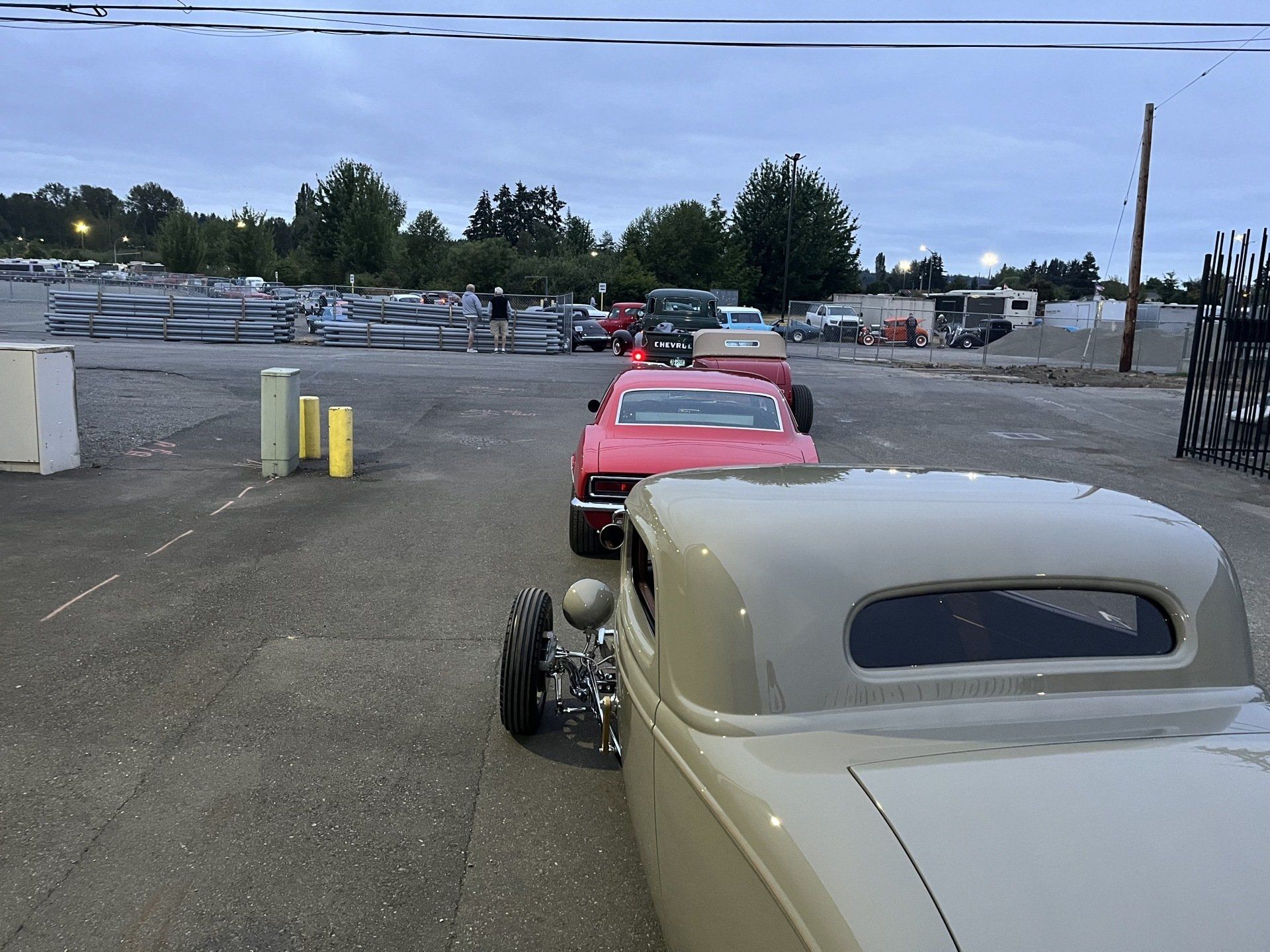 Two vintage hot rods parked in a lot, one cream-colored in the foreground and a red one behind it.