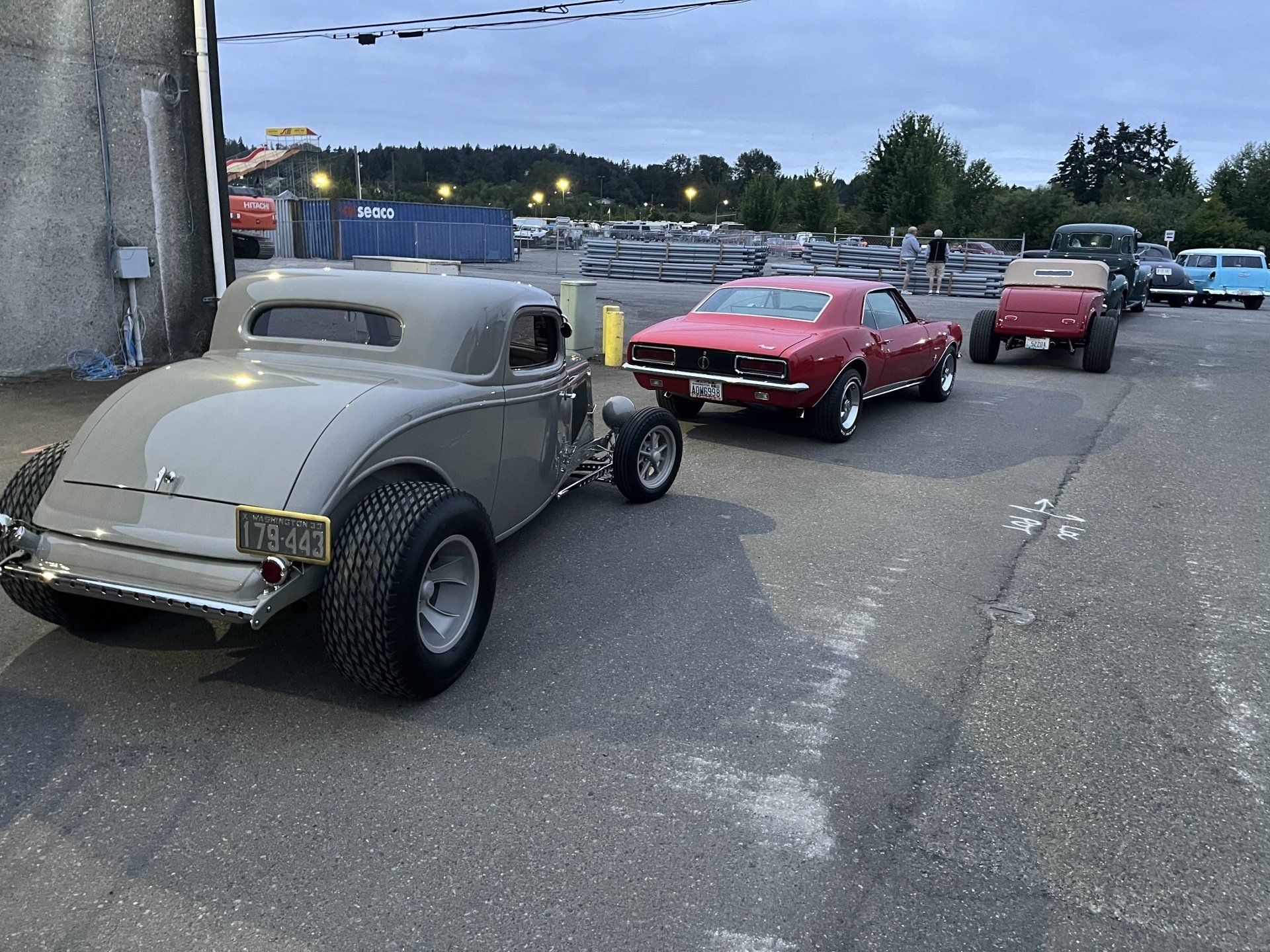 Vintage cars lined up in a parking lot at dusk, including a gray hot rod and red classic coupes.