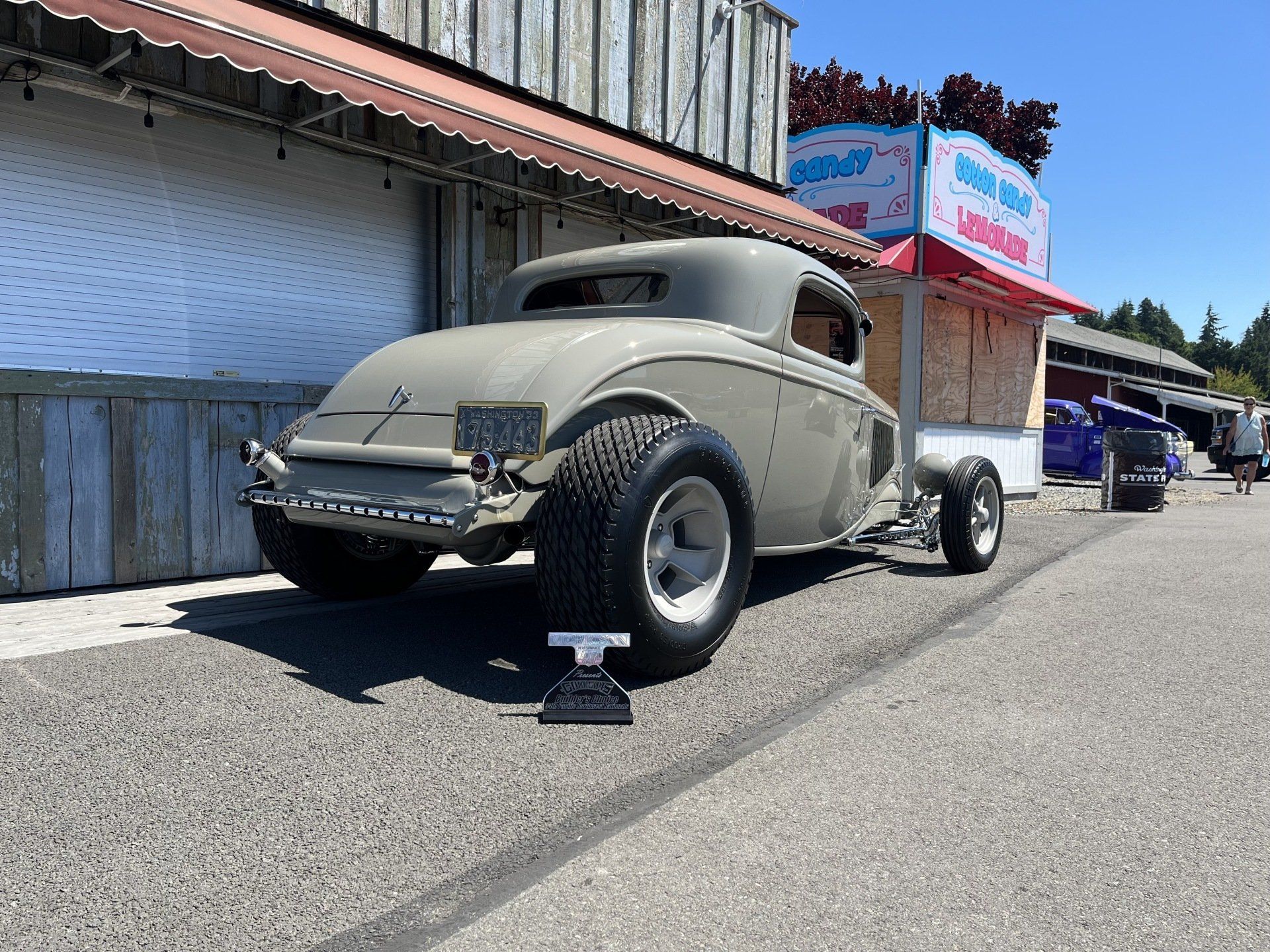 Vintage beige hot rod parked on a street beside a building at an outdoor car show.