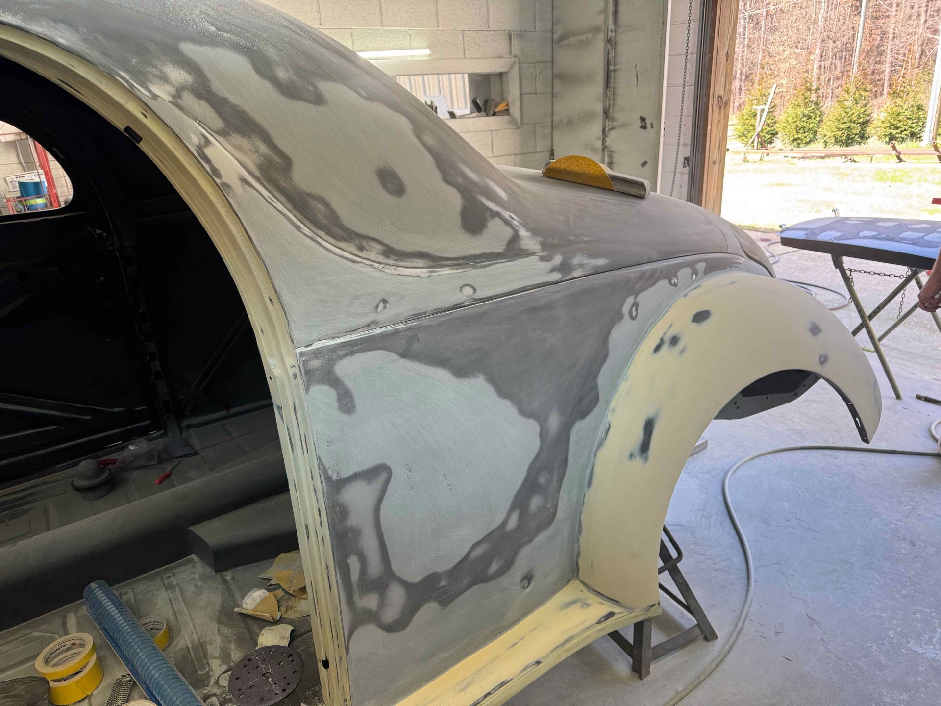 Car body panel being sanded and primed in a garage, with gray body filler patches visible.