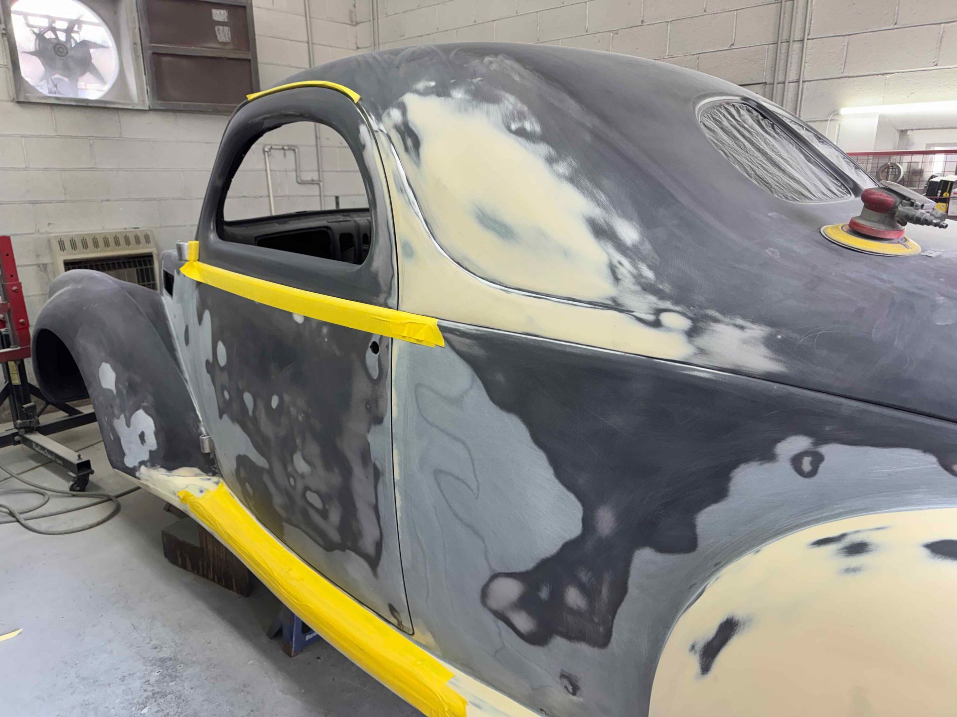 Partially painted black coupe in a body shop, with yellow masking tape and primer patches visible