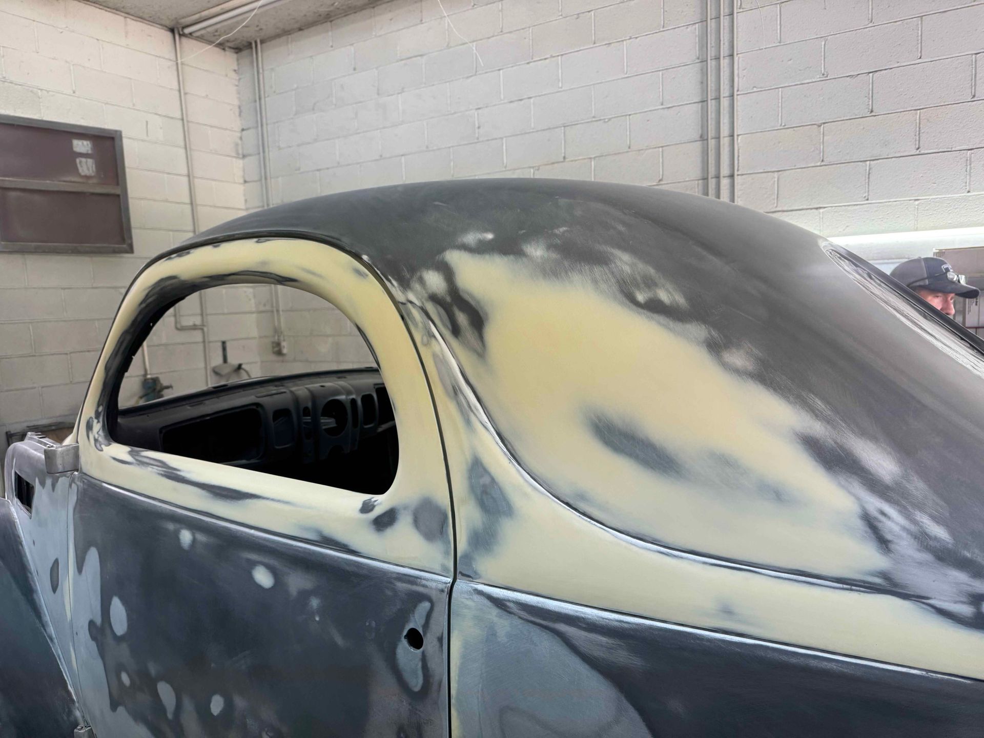 Primered classic car body in a workshop, with sanded gray and yellow patches.