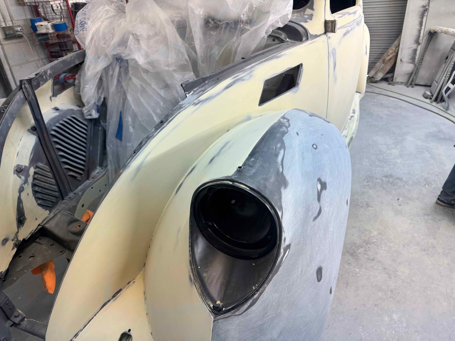 Classic car body in workshop, with a large gray headlight housing and cream-colored paneling under repair