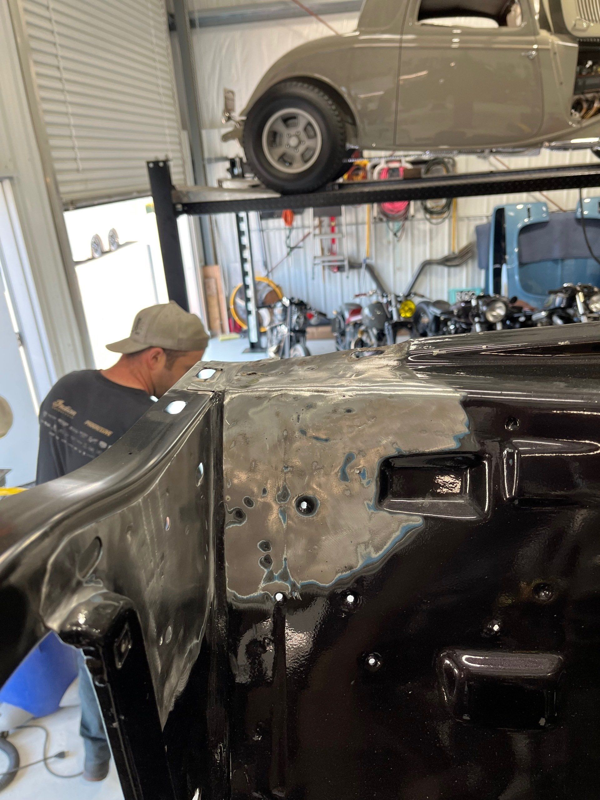 Person working on a black car in a garage, with another car on a lift above.