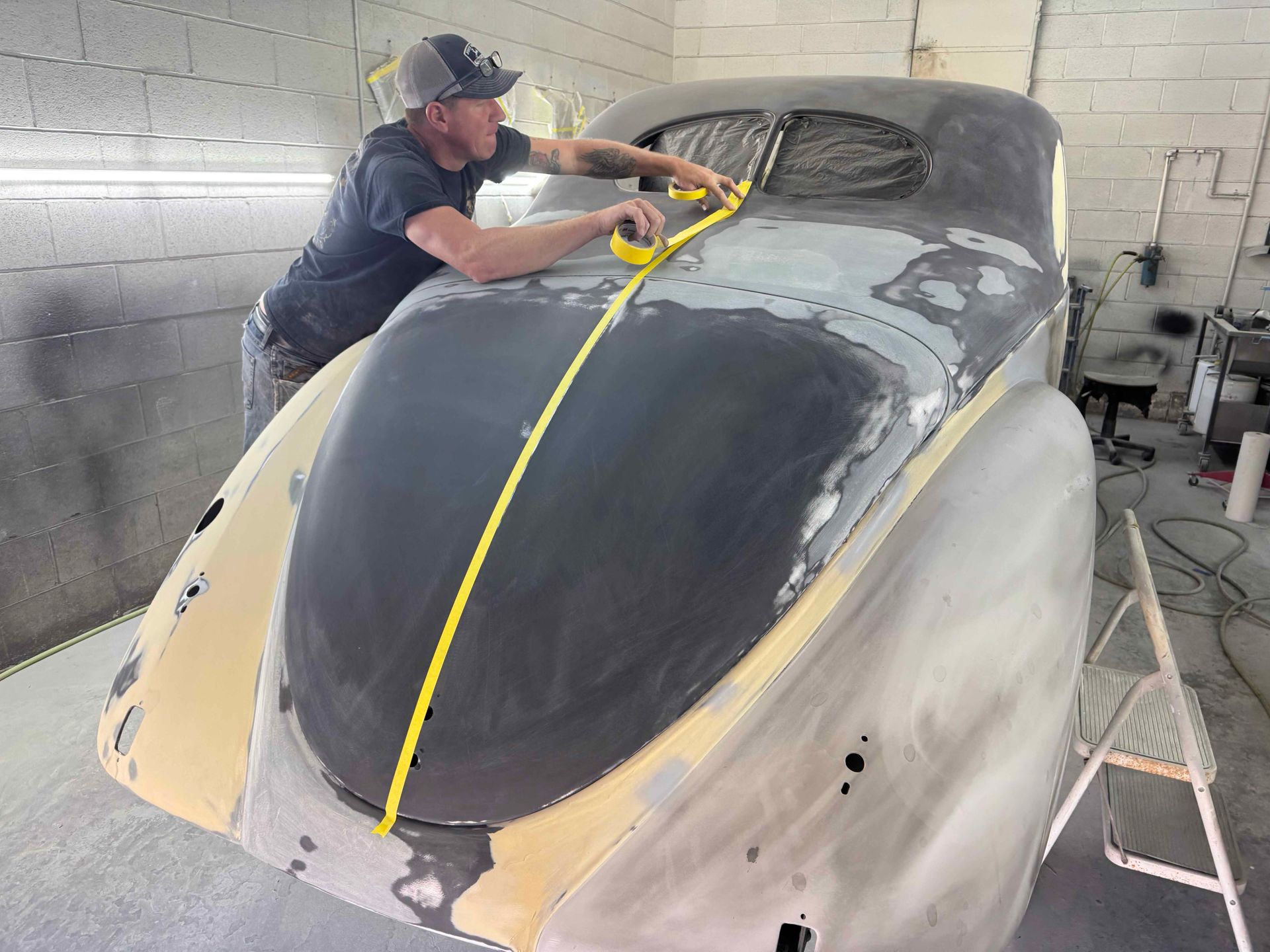Worker sanding a car body in a garage, with yellow masking tape on the hood.