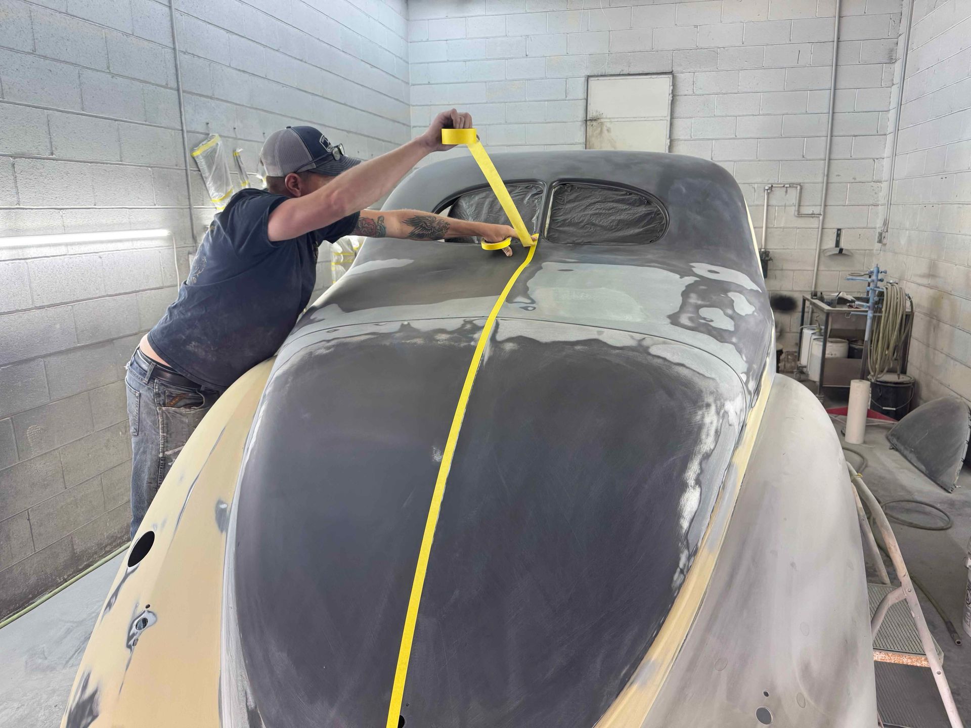 Person sanding the nose of a vintage car in a workshop, with yellow masking tape down the center.