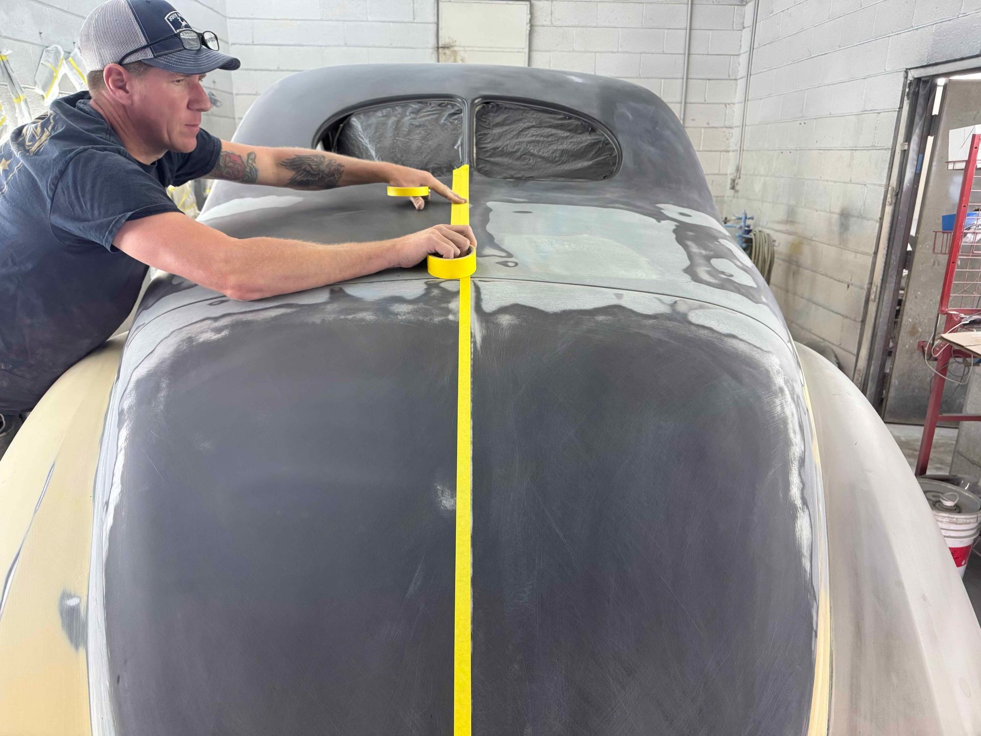 Technician measuring and sanding a vintage car body in a body shop with a yellow tape line down the center