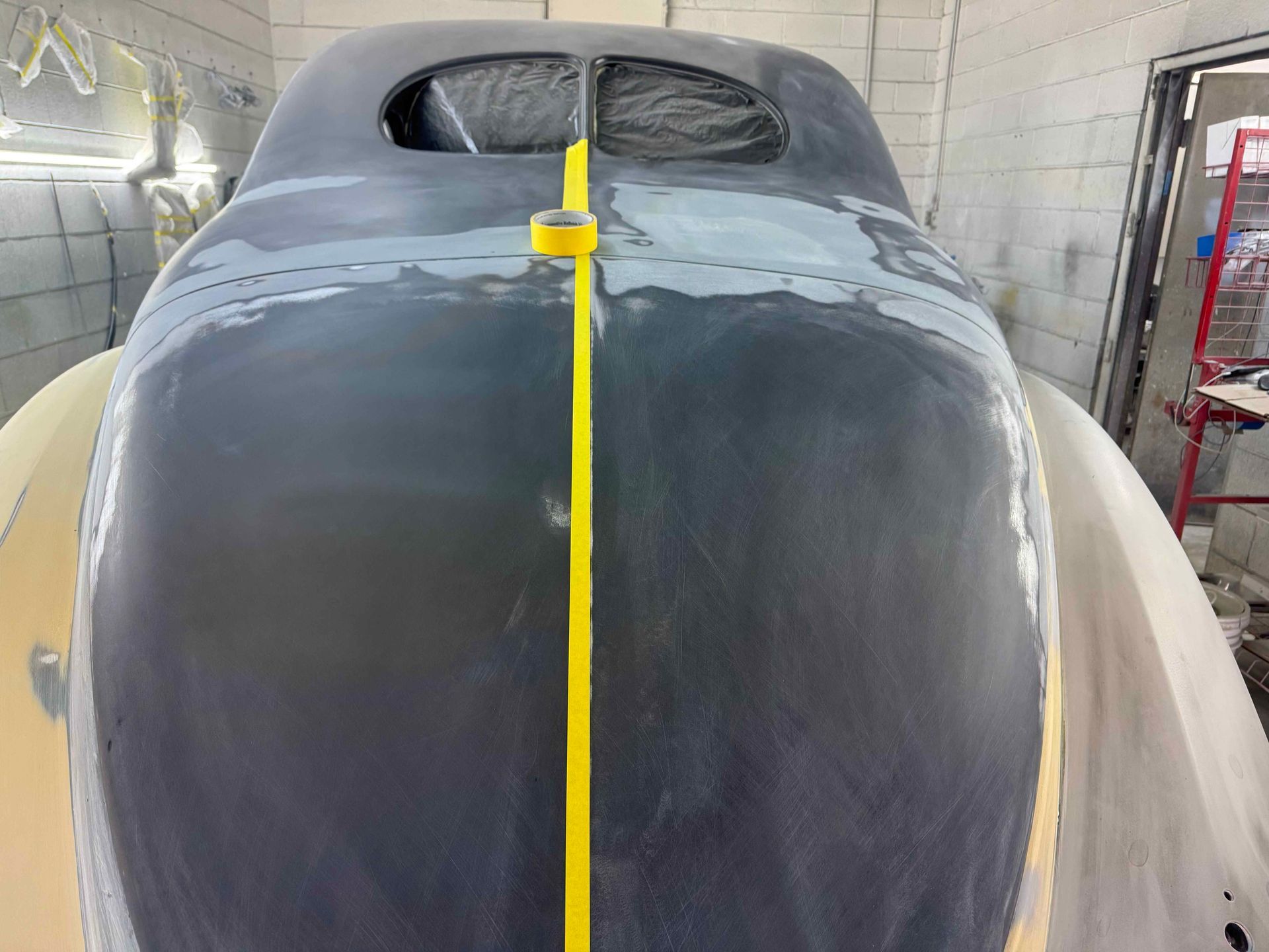 Front view of a gray car body in a paint booth, with a yellow tape line down the center.