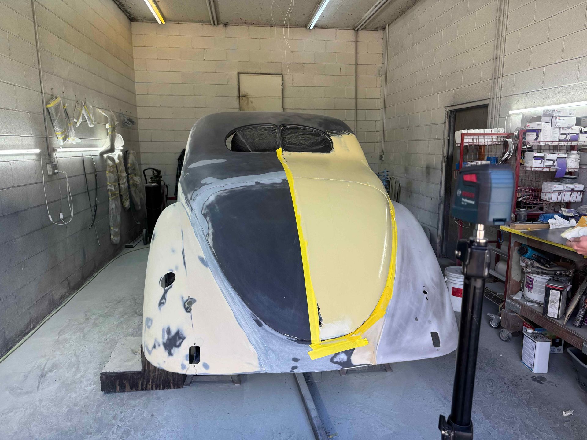 Partially painted vintage car in a garage, with gray primer, yellow masking tape, and white bodywork surfaces