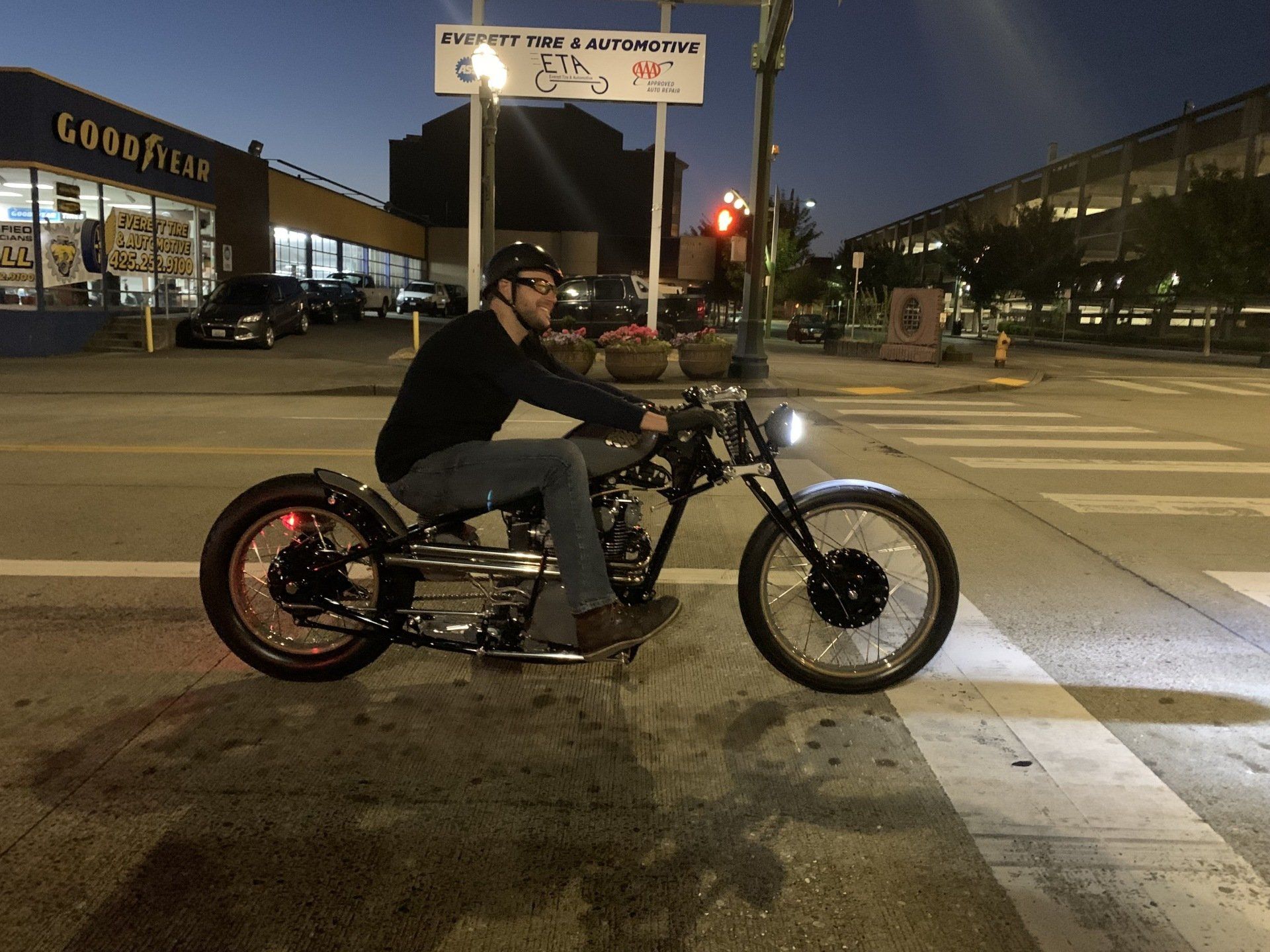 Man riding a custom motorcycle at night on a city street, with lit storefronts in the background.