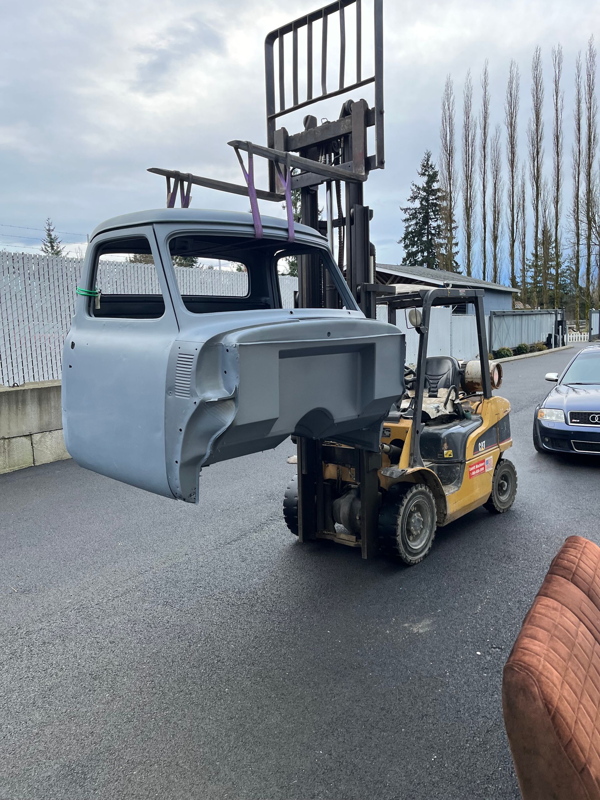Yellow forklift lifting a gray truck cab in a gravel yard