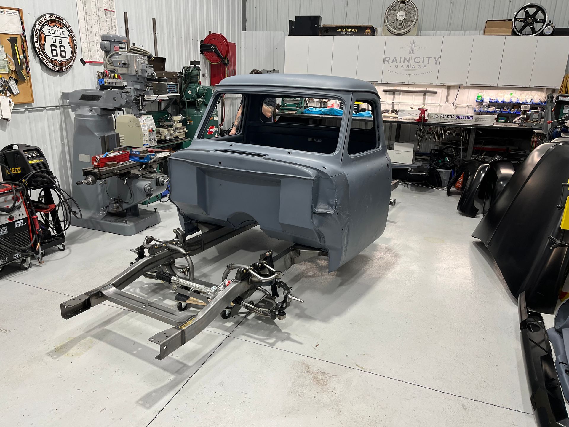 Gray classic truck cab on a chassis in a workshop, surrounded by auto parts and tools.