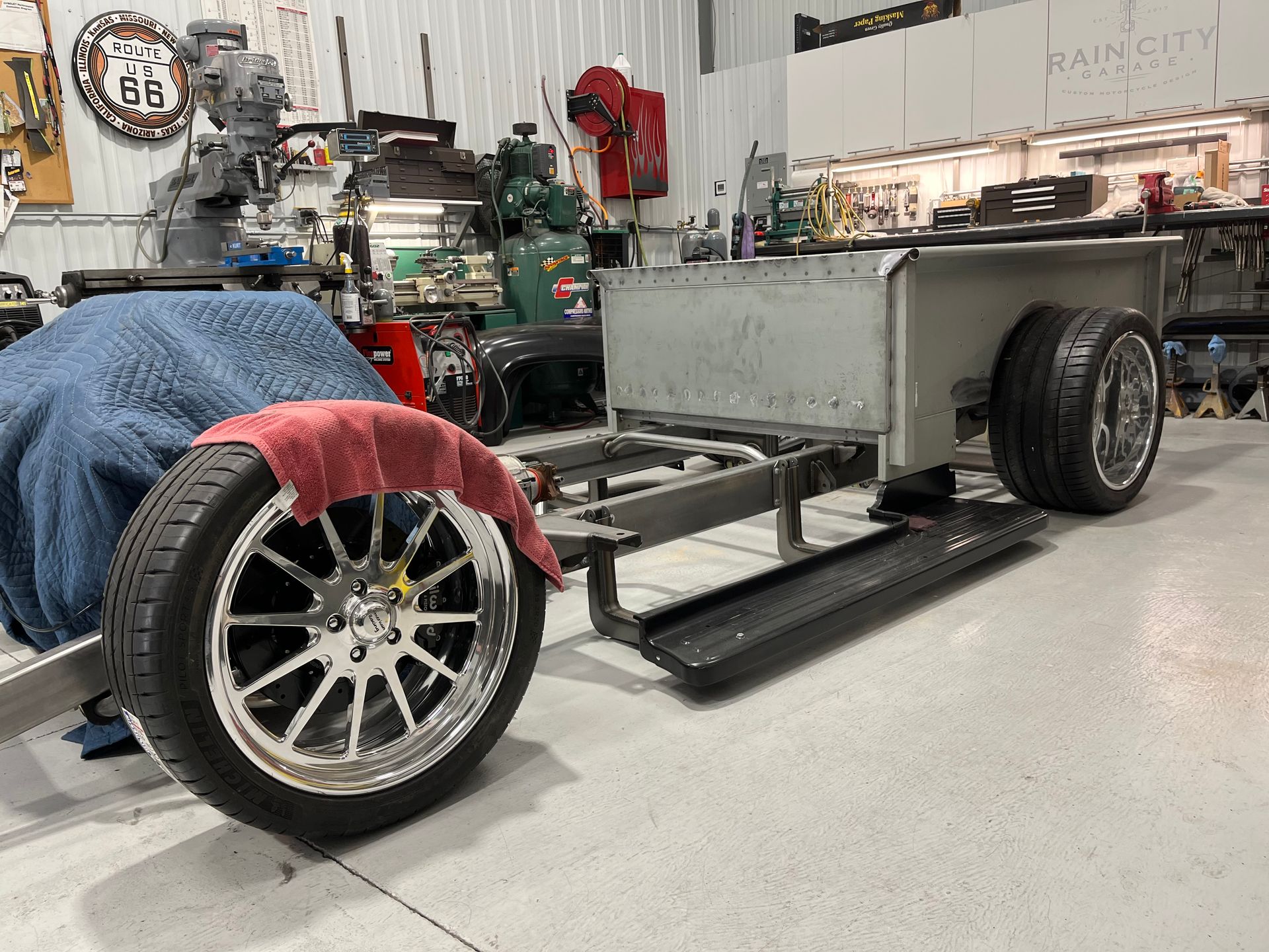 Custom car frame in a garage, with large wheels and colored blankets draped over the chassis.