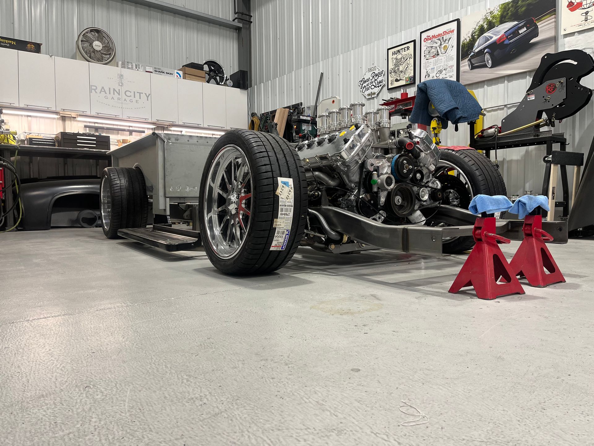 Classic car chassis in a workshop, with a mechanic working on the engine and red jack stands in front.