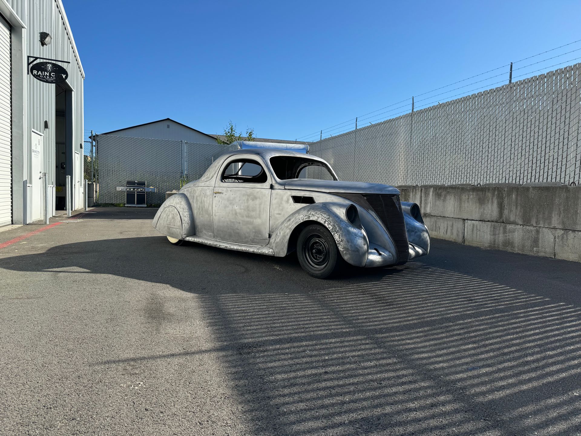 Vintage gray car parked in an industrial lot beside buildings and a chain-link fence.