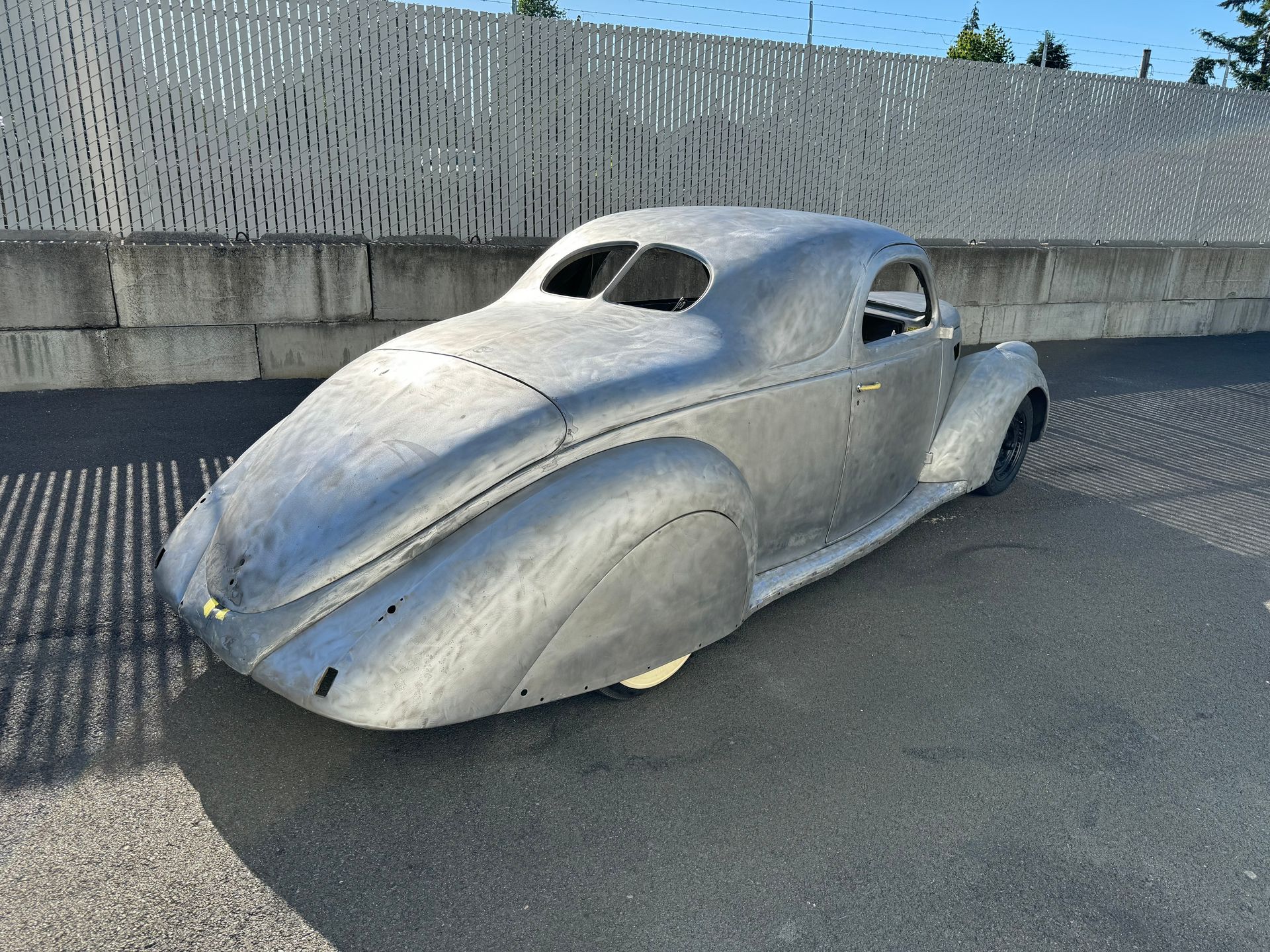 Silver vintage hot rod body with a low, streamlined design parked outdoors on pavement.
