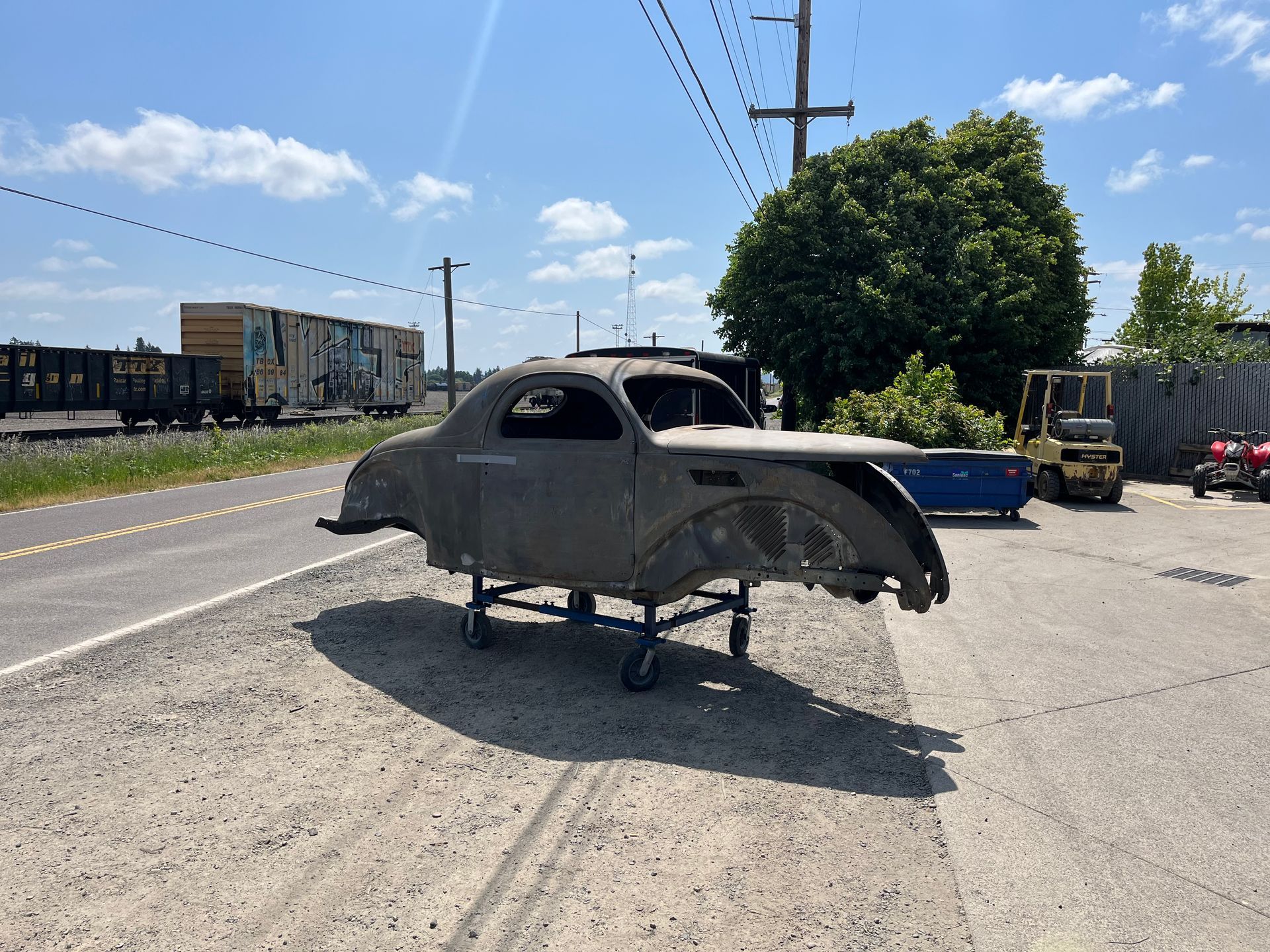 A gray, unfinished car body sits on jack stands in a roadside lot under a bright blue sky.