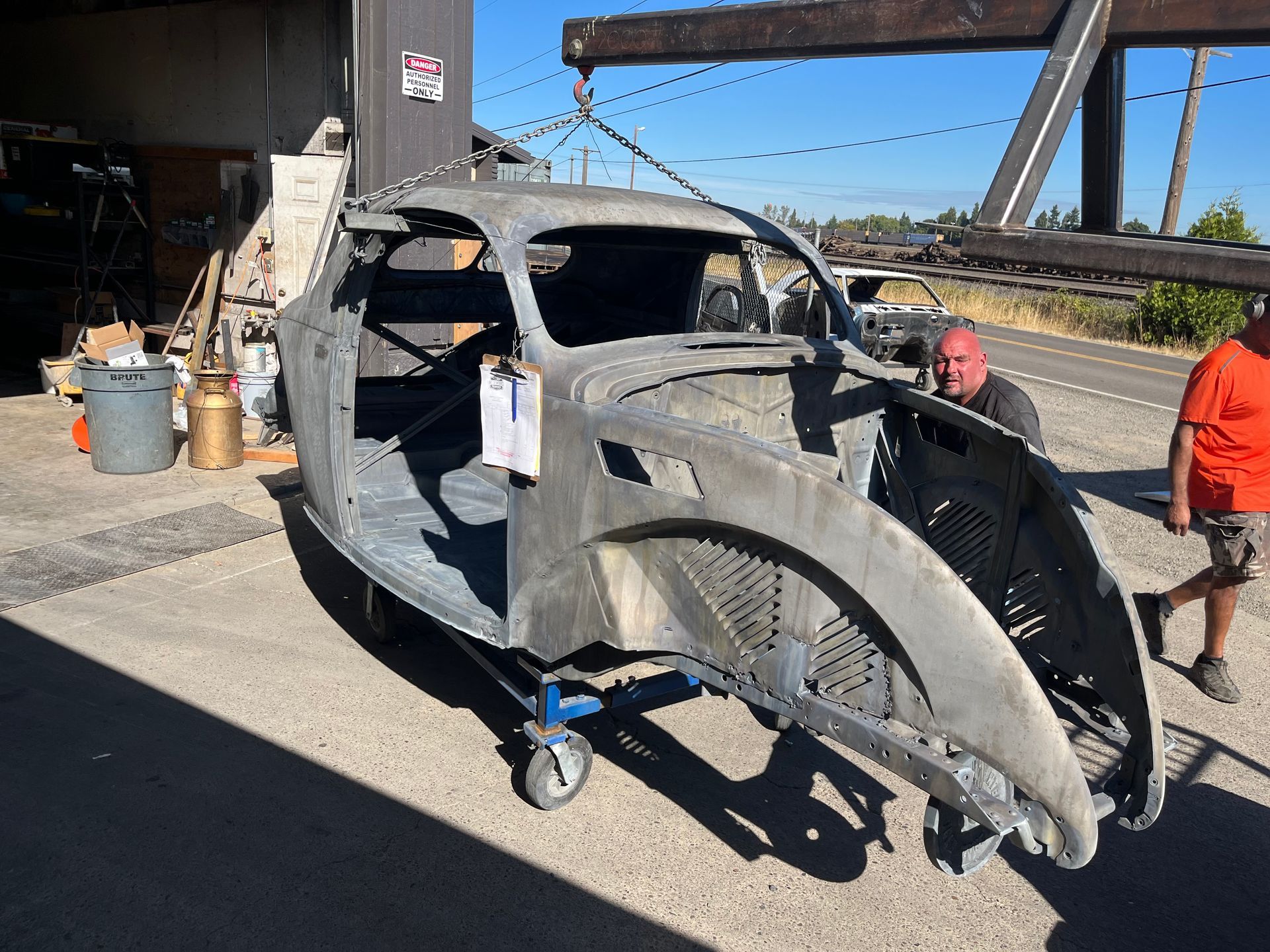 Damaged vintage car body on a wheeled dolly, with two workers in an industrial yard, gray primer finish
