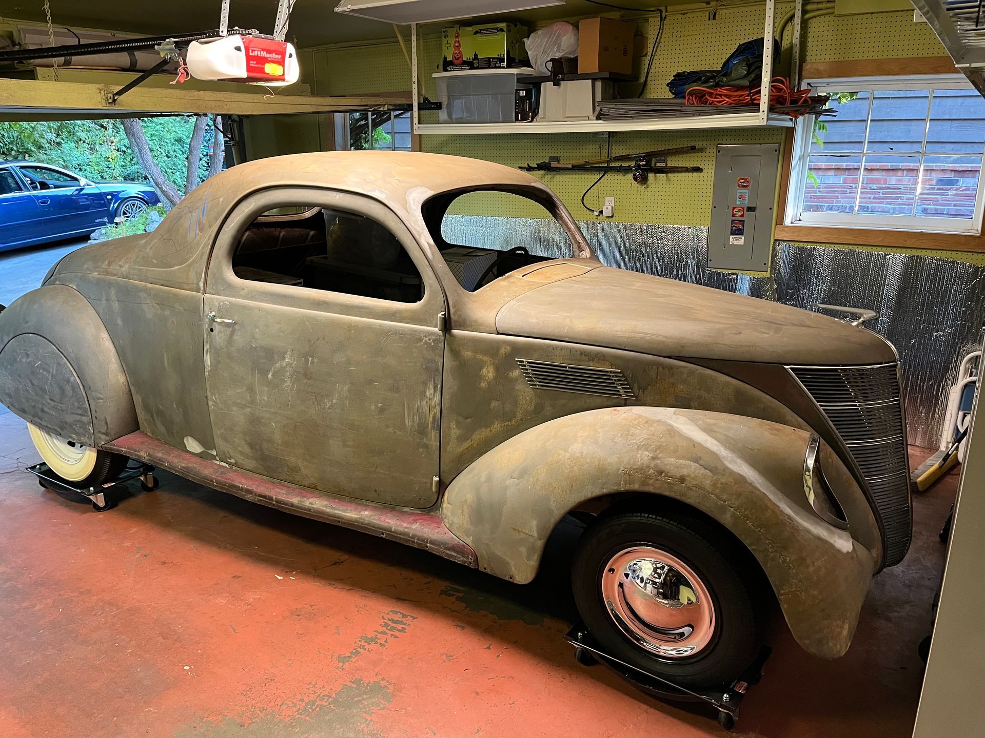Dusty vintage coupe in a garage, with rounded fenders and an unfinished olive-green body