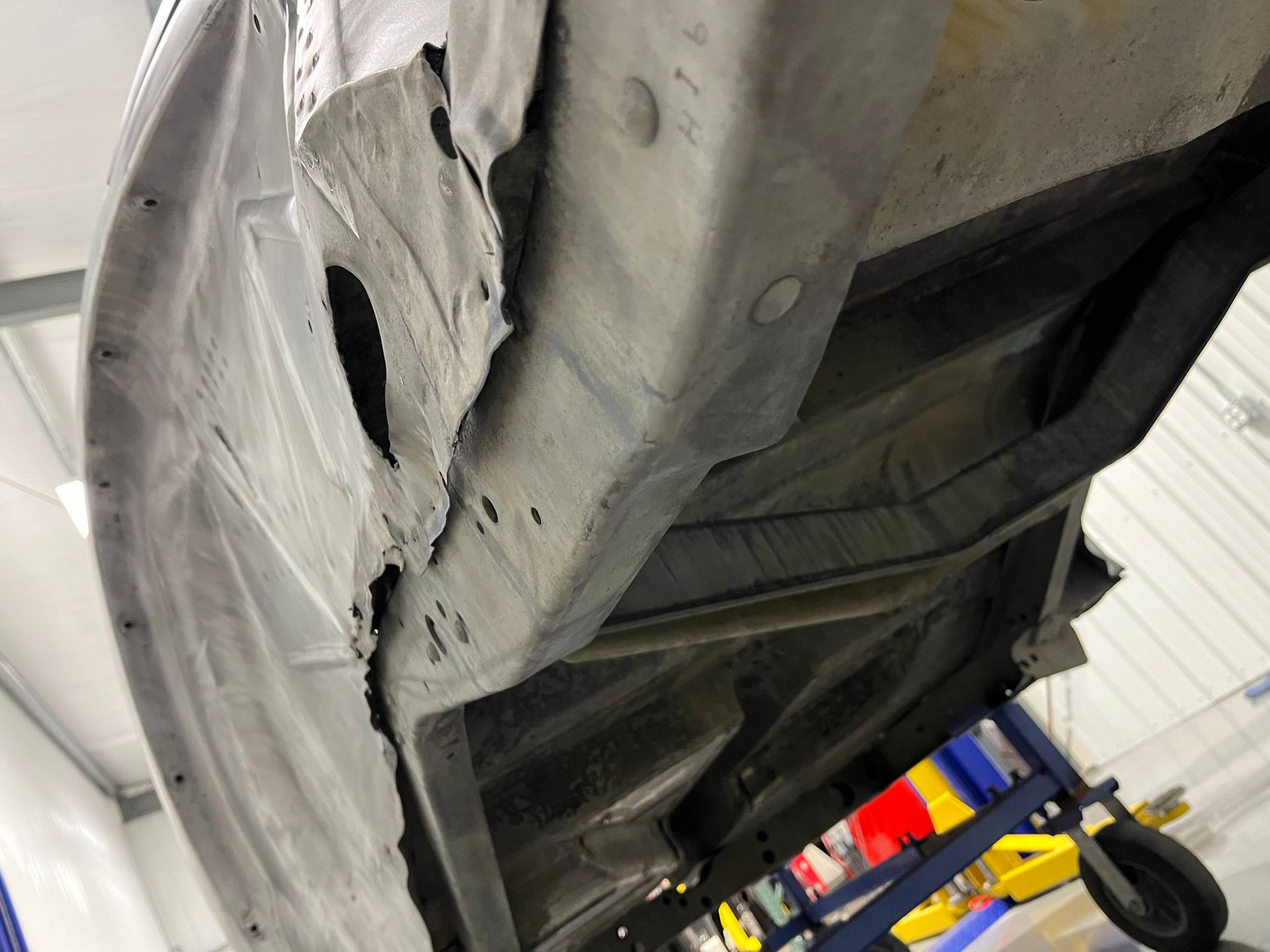 Undercarriage of a damaged car on a lift, with torn metal paneling and a wheel visible in the background.