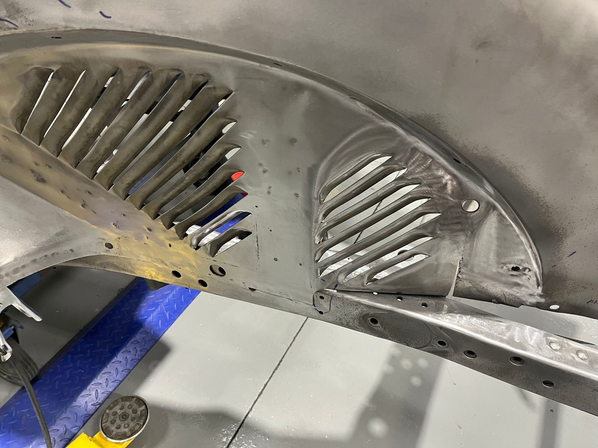 Curved metal panel with slotted vents and rivets on a workshop floor, with a blue jack support nearby
