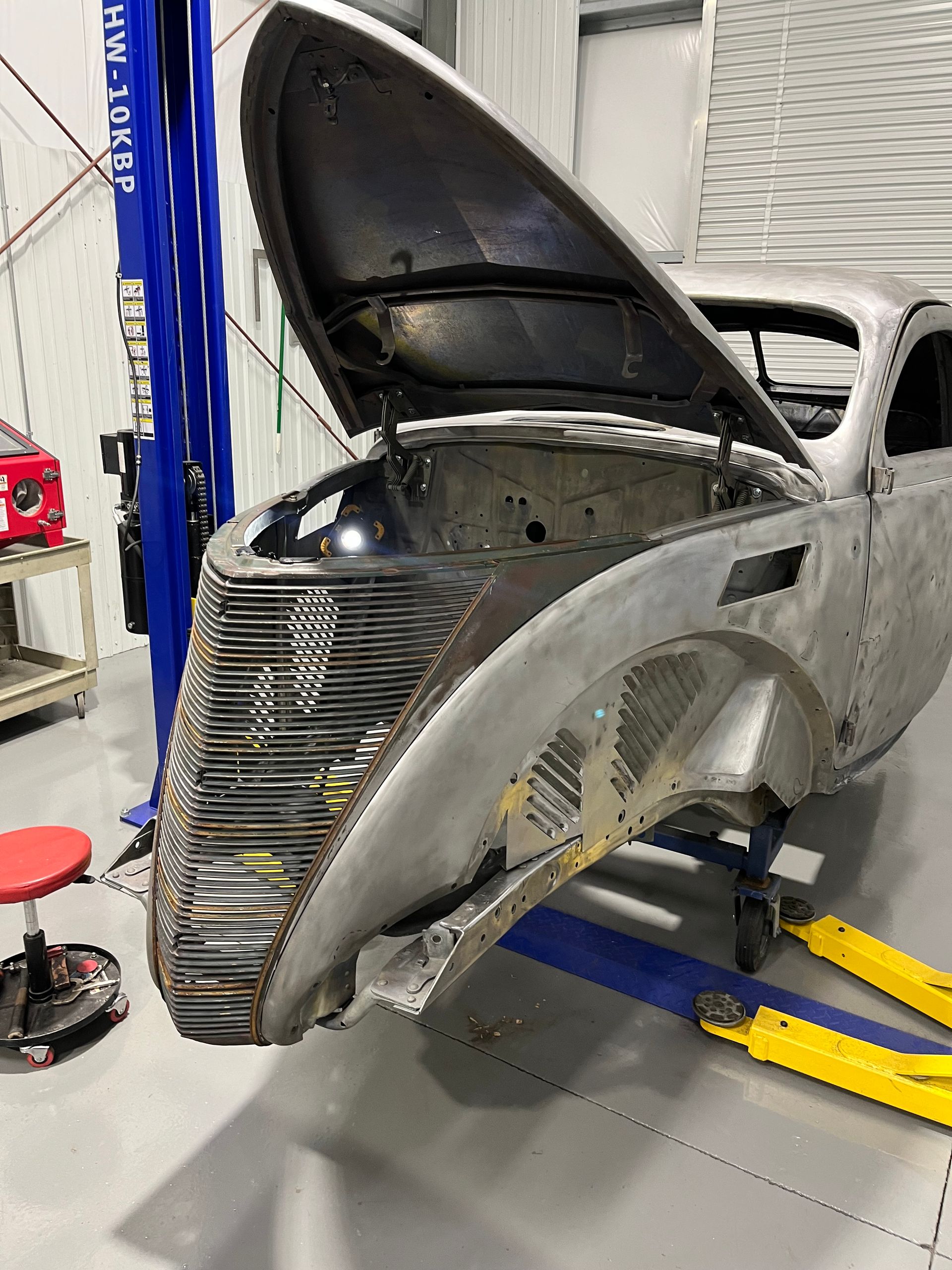 Car body in auto repair shop with hood open, showing front end and exposed interior frame.