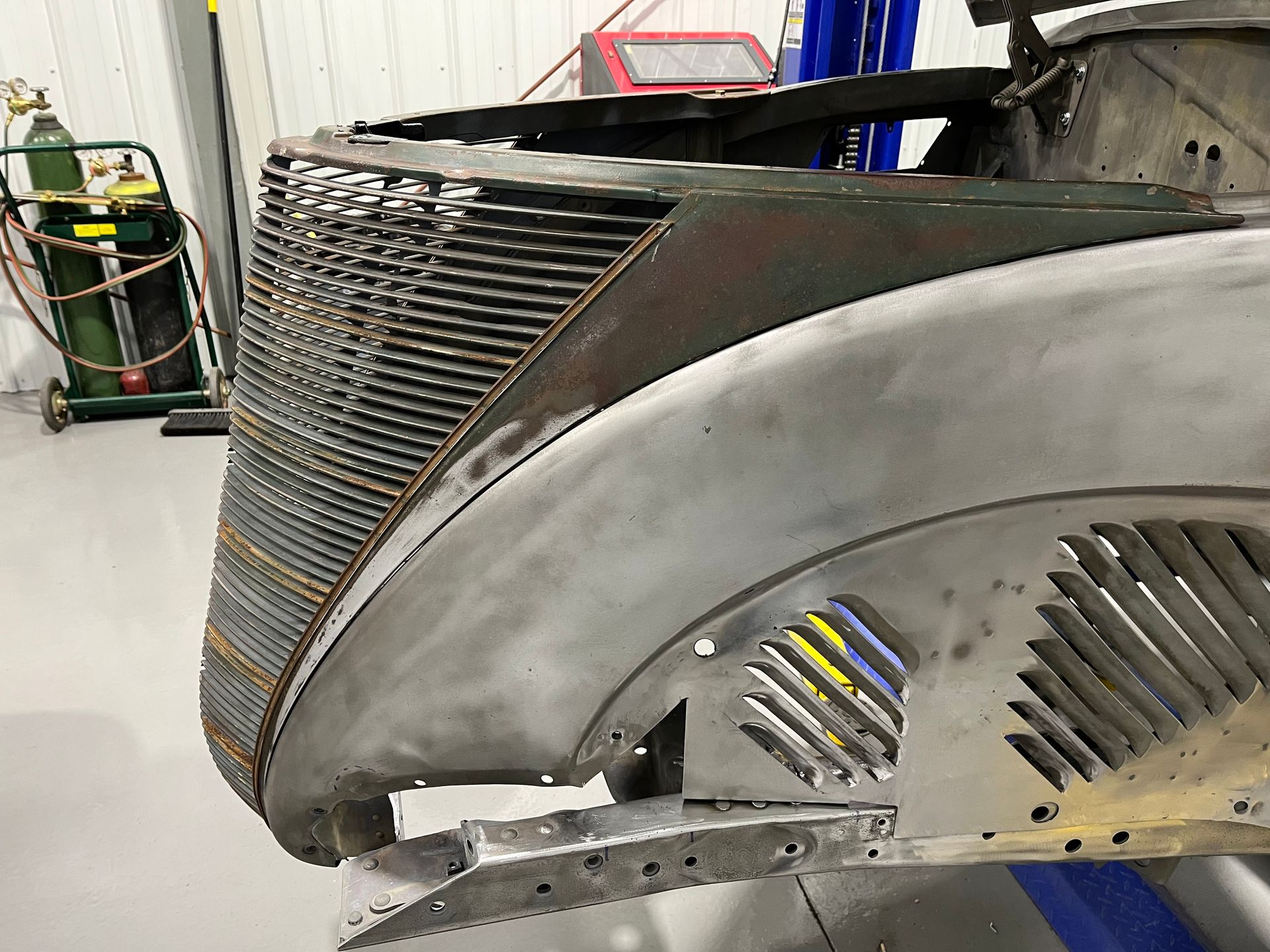 Close-up of a partially disassembled silver vehicle body in a workshop, showing a slatted grille and side vents.