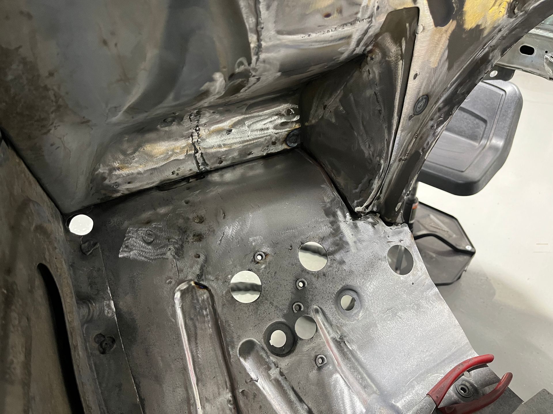 Damaged metal floor with rust, drilled holes, and a partially visible red tool near a vehicle seat base