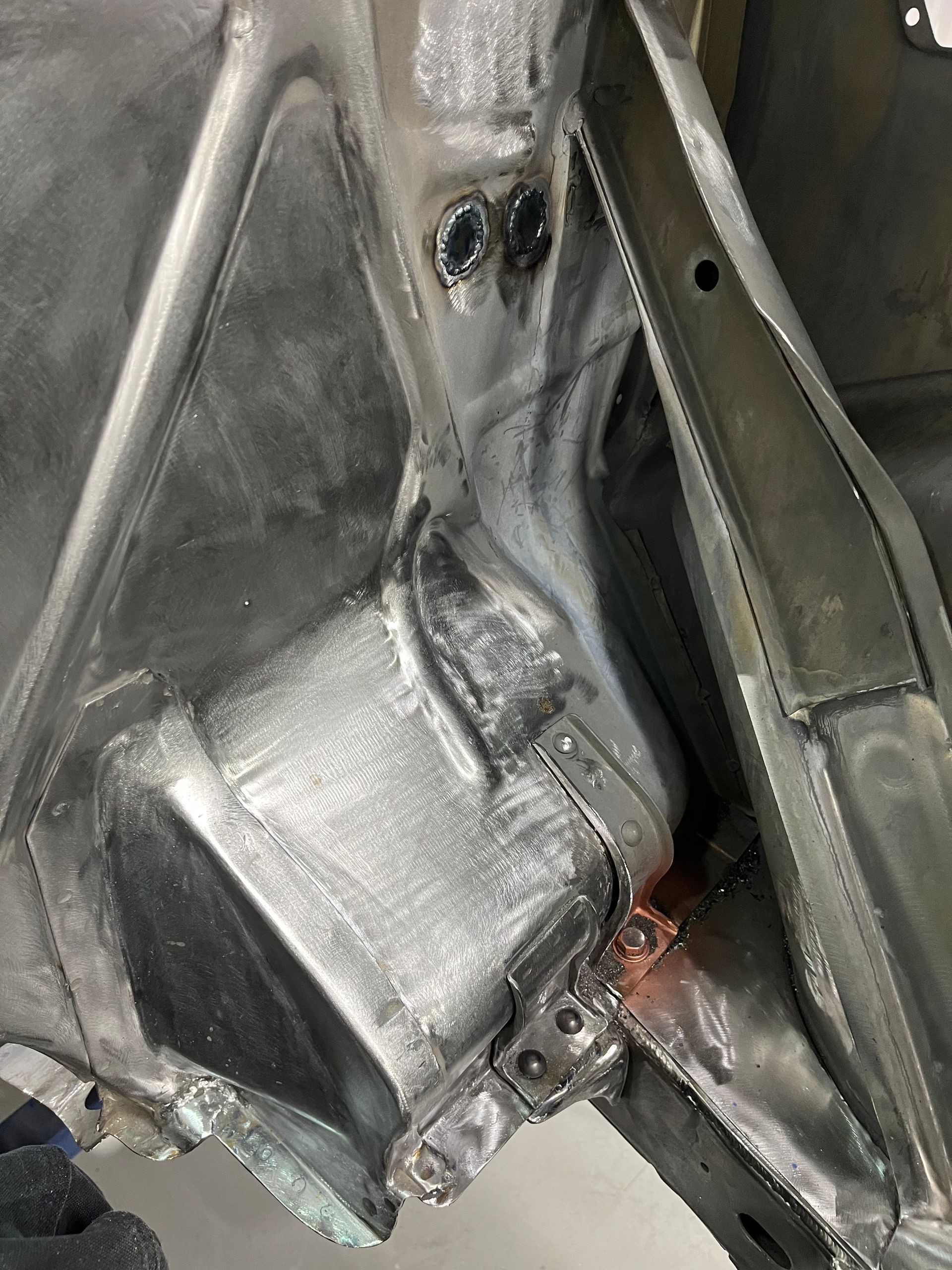 Close-up of a metallic car chassis with welded seams and structural braces, showing a silver engine or component mount.