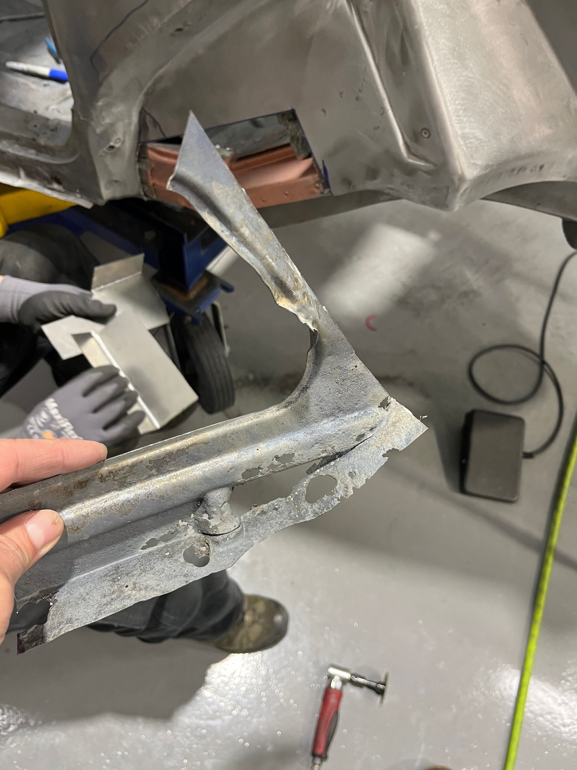 Damaged metal car part being held in a workshop, with tools and floor visible.