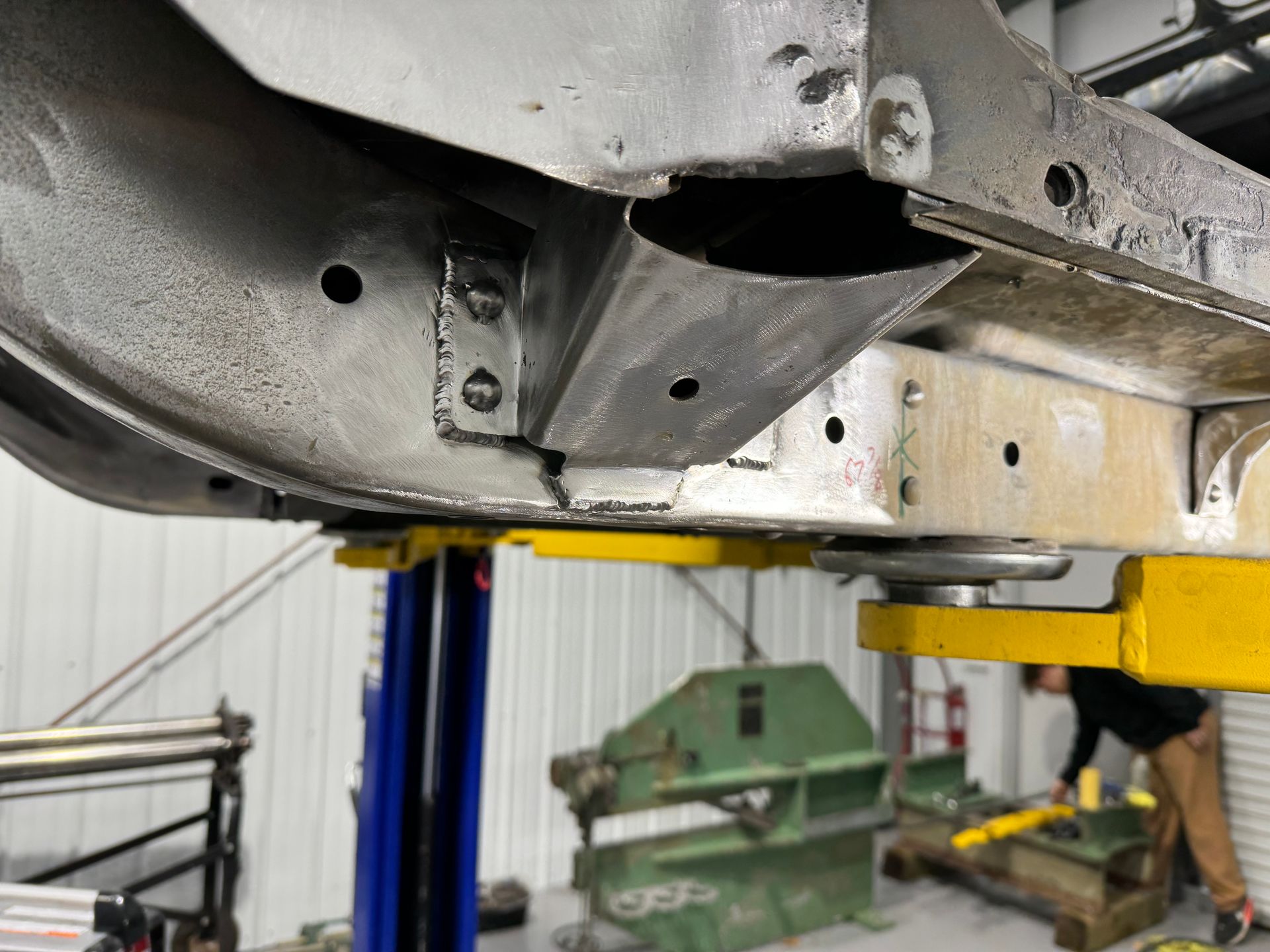 Undercarriage of a silver vehicle on a lift in an auto repair shop, with a mechanic nearby