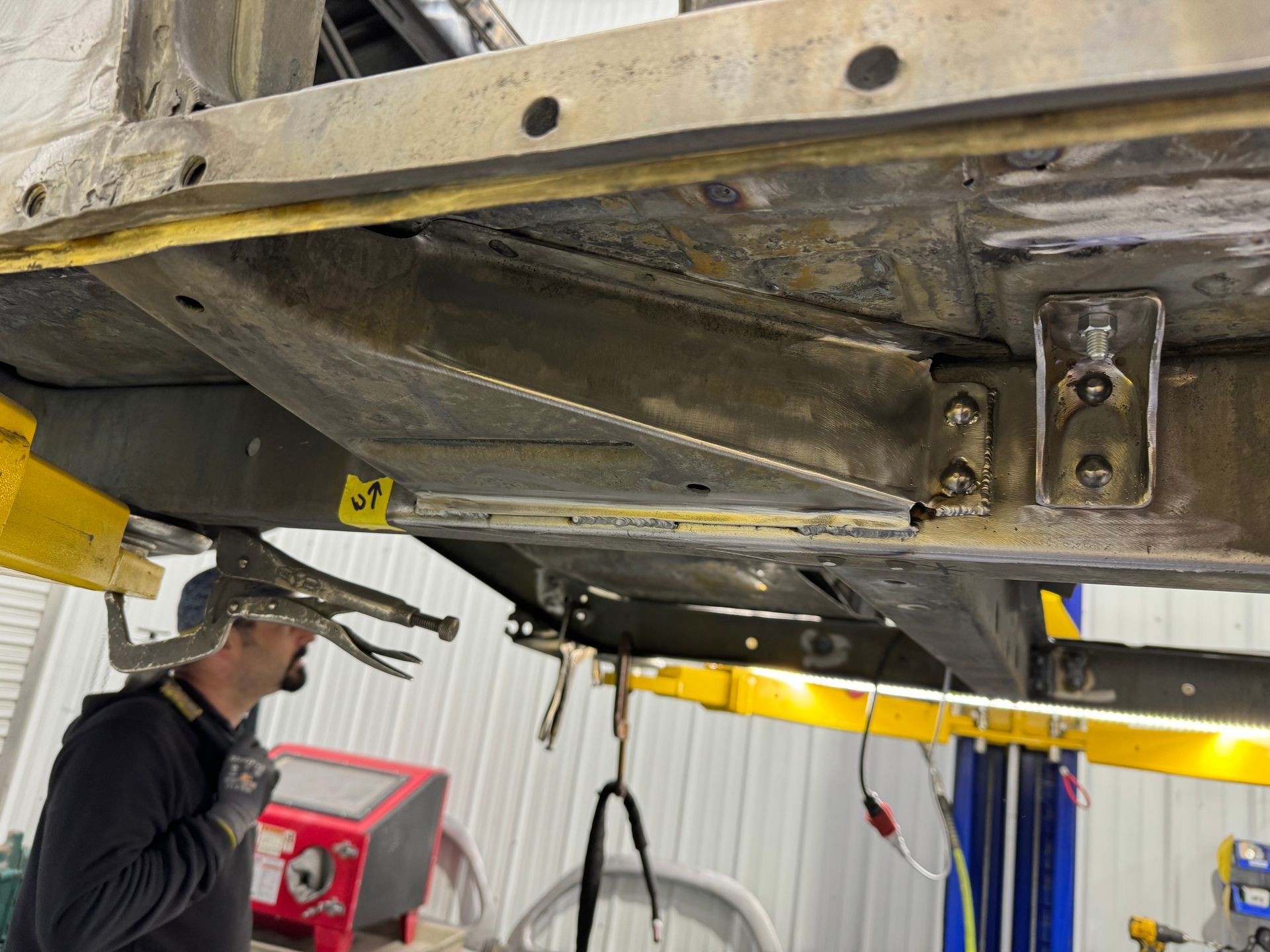 Mechanic inspecting vehicle undercarriage on a lift in a garage, with tools and yellow lift arms visible