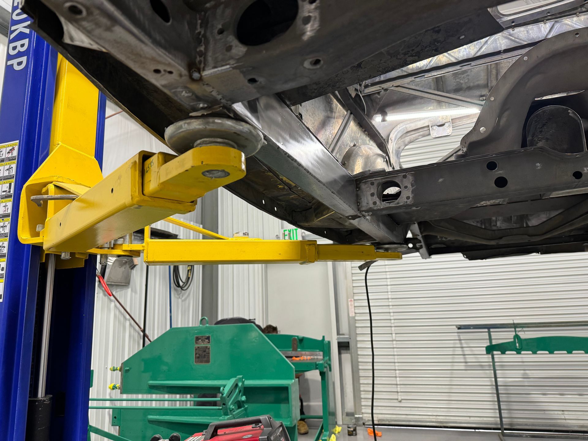 Yellow lift arm supporting a car underside in an auto shop beside green trash bins