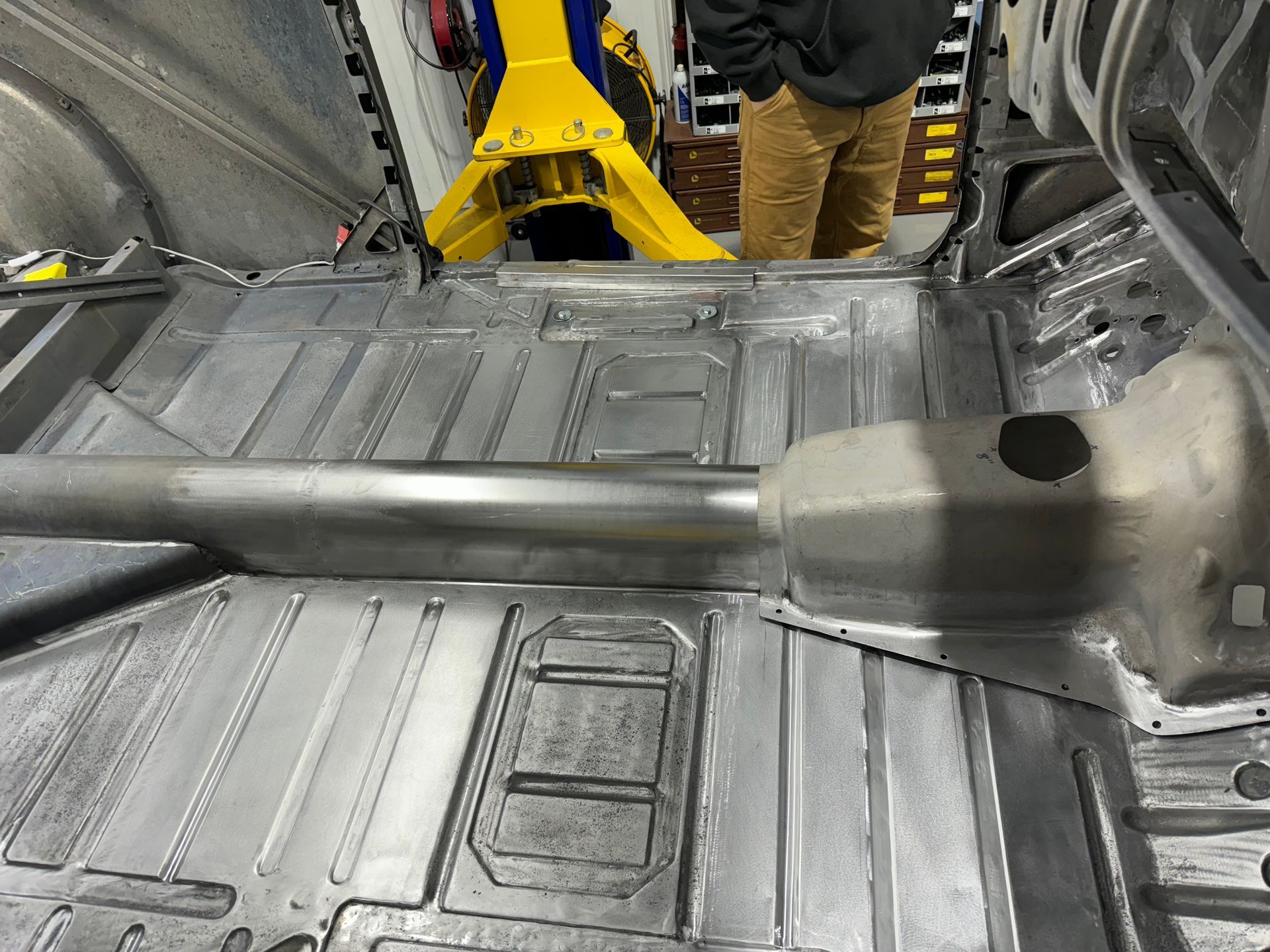 Metal car interior floor with a long silver exhaust or tunnel component being installed by a worker in brown boots.