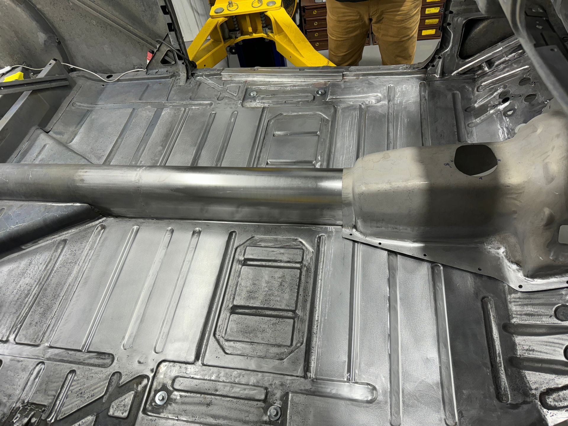 Metal driveshaft on a car floor pan in a repair shop, with a worker visible in the background.