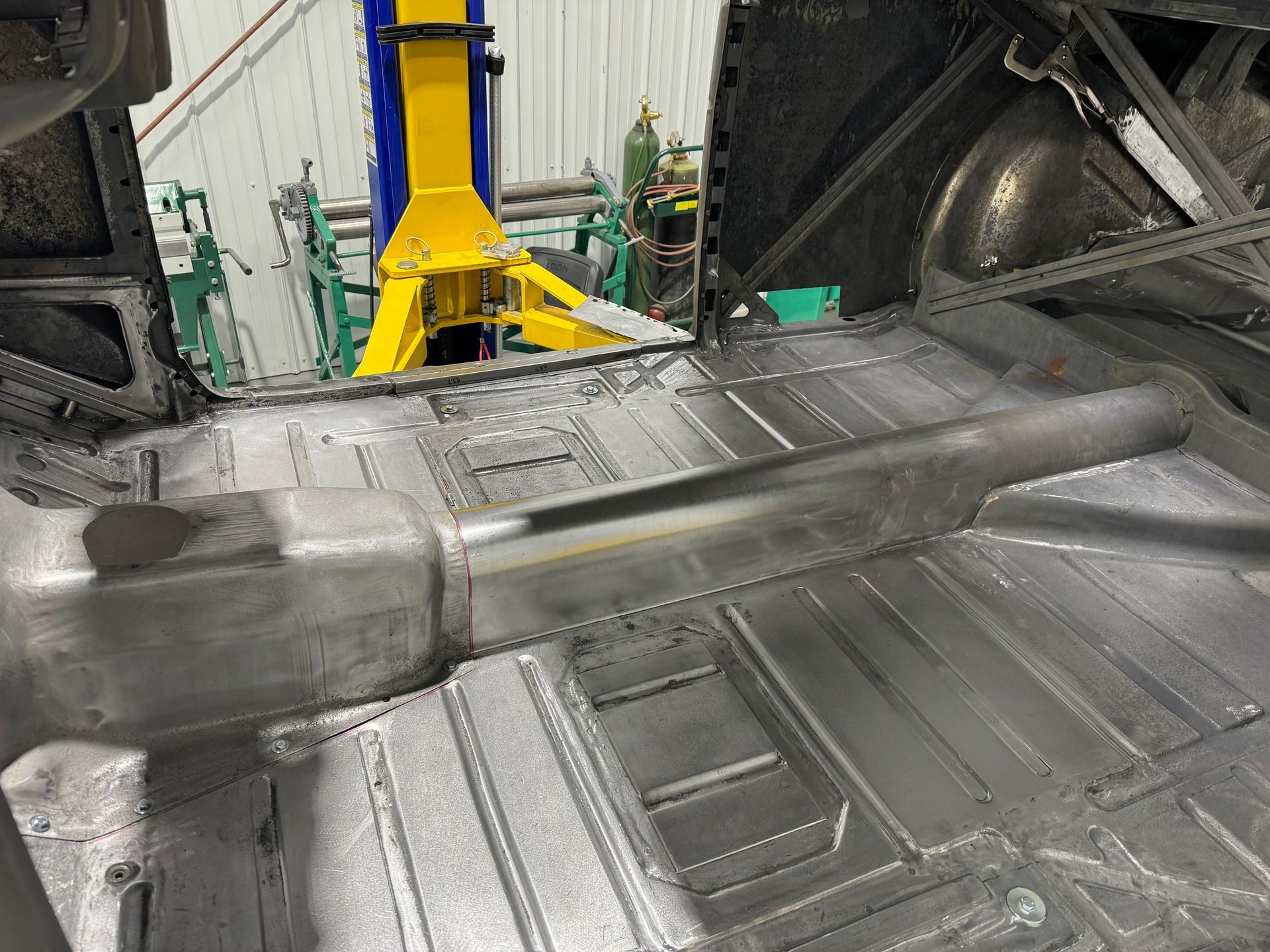 Inside a car factory, a bare metal vehicle floor sits on an assembly jig with yellow equipment nearby.