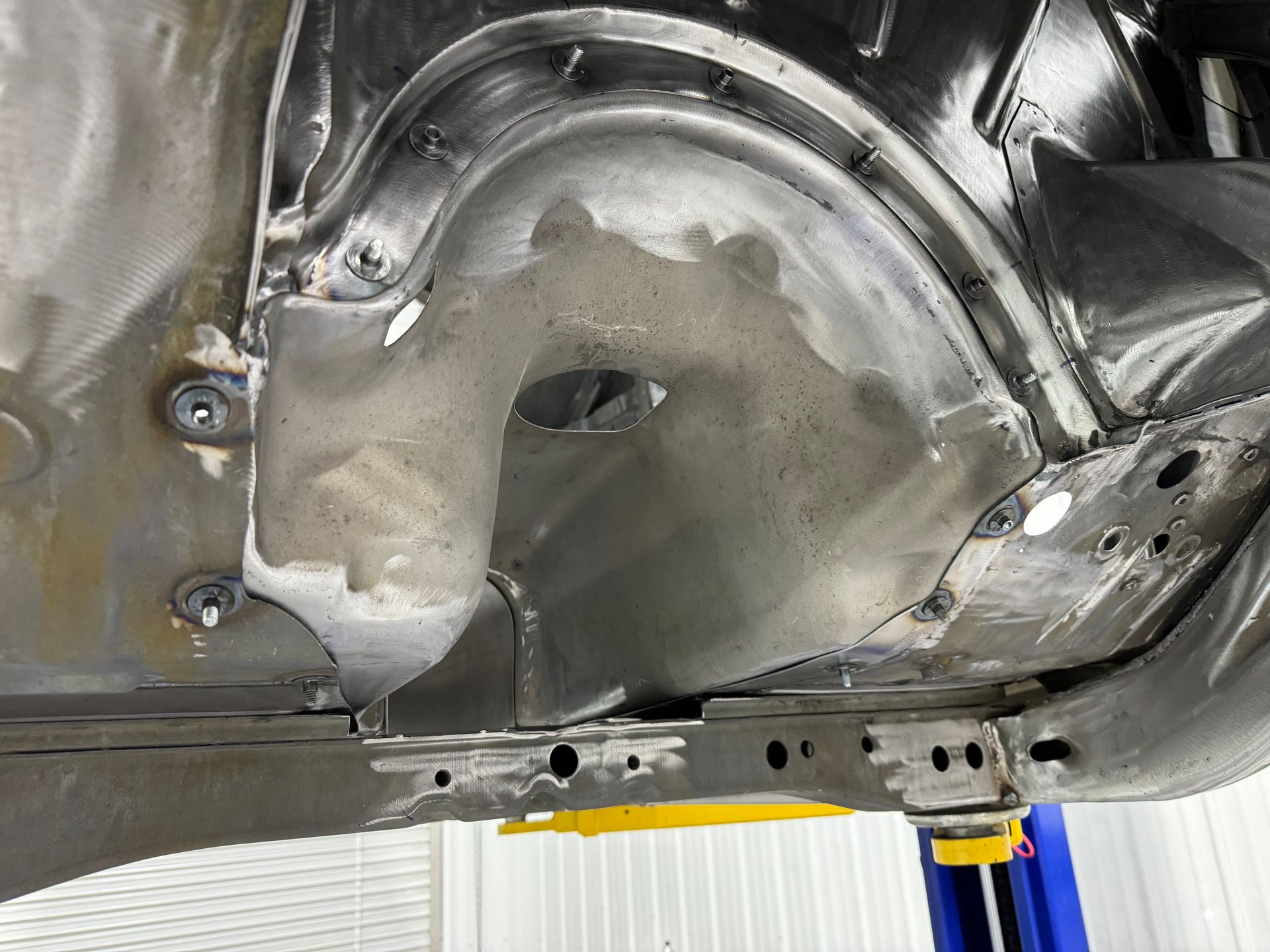 Close-up of a car’s metal underbody with rust spots, holes, and a yellow jack stand below