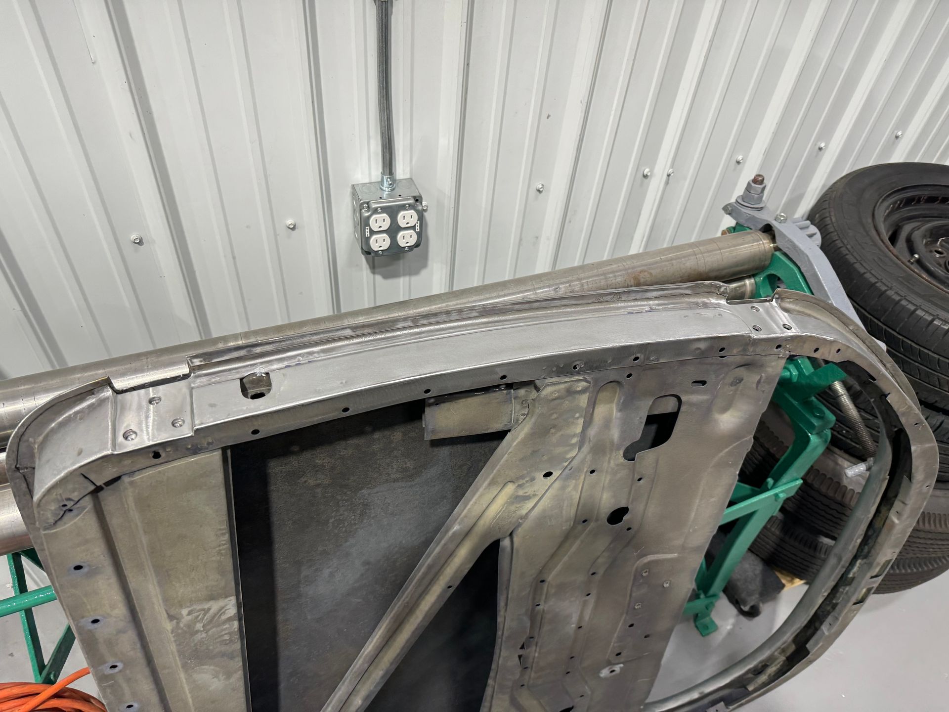 Upside-down car body frame in a garage, with exposed metal, wires, and a spare tire nearby