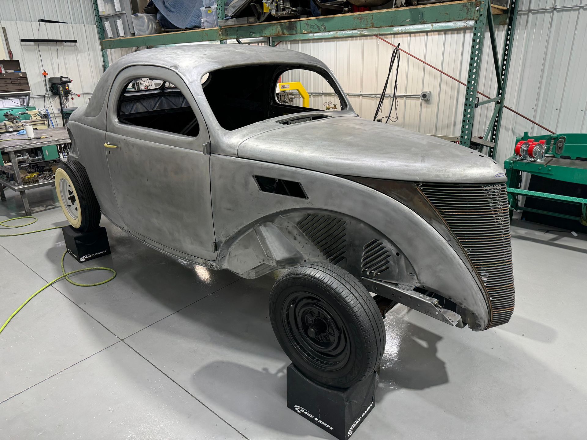 Gray vintage car body shell on stands in a workshop, unfinished with no windows or paint.
