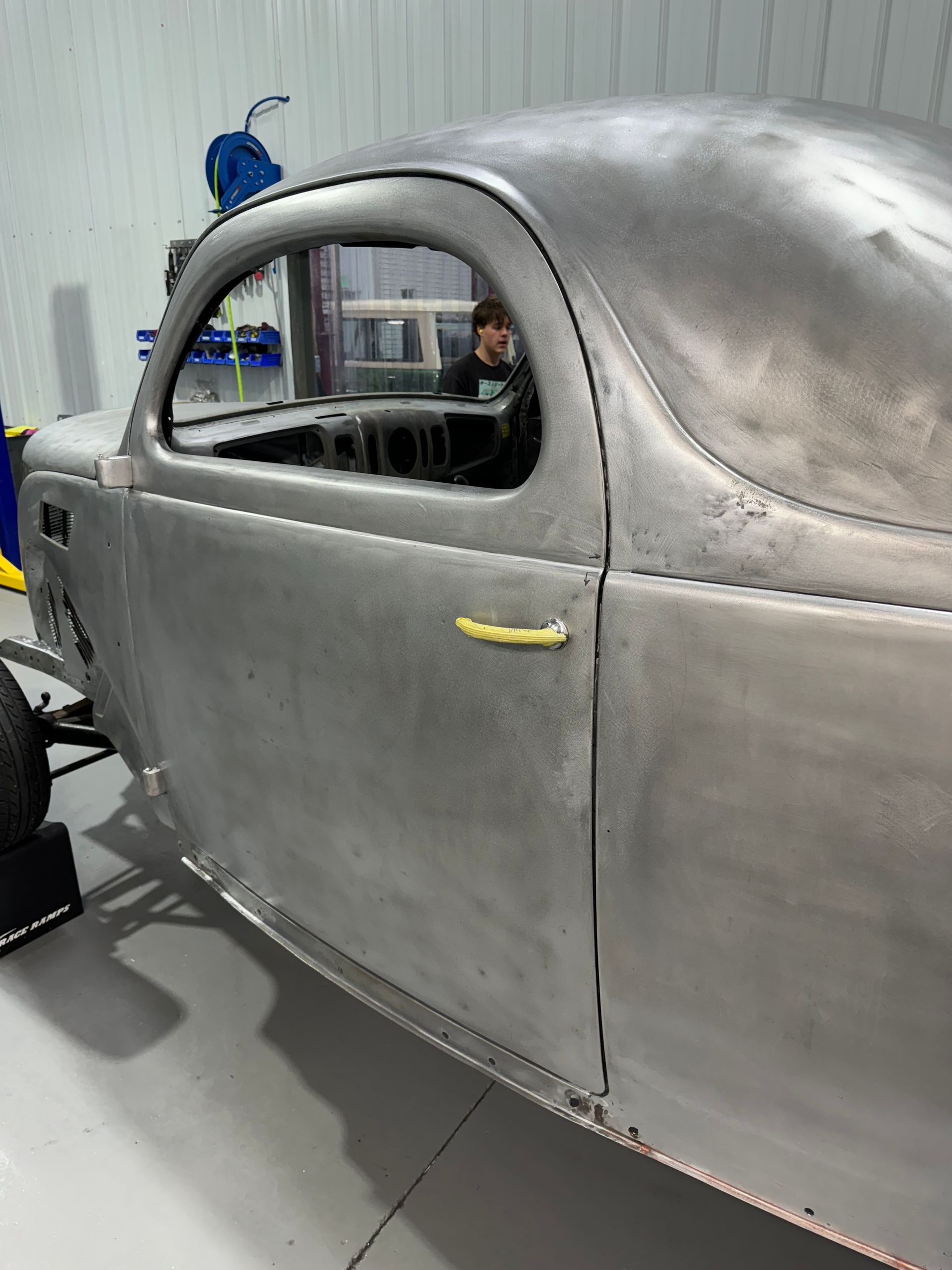 Gray vintage car body in a workshop, with a blue helmet visible through the window