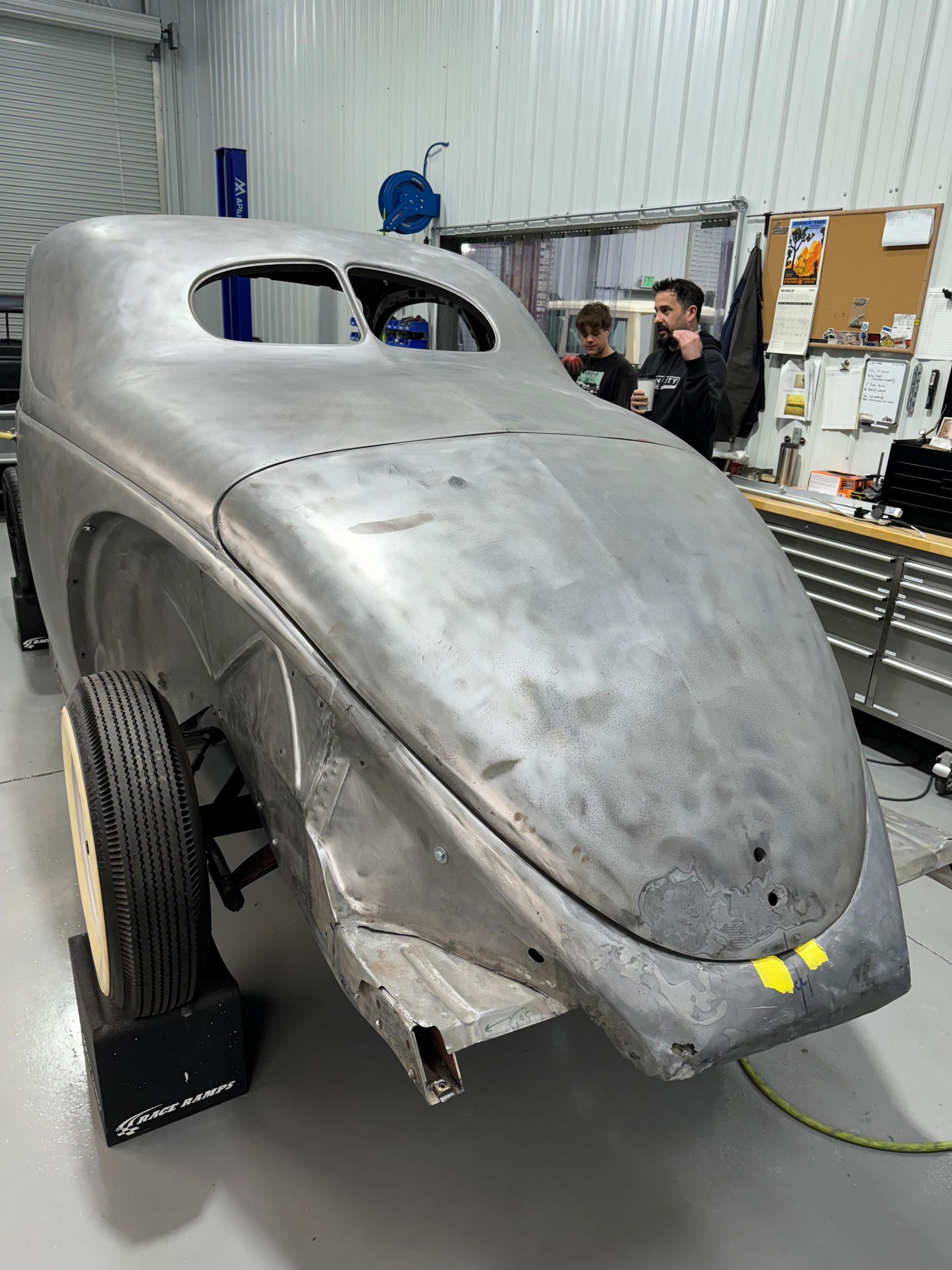Silver vintage car body in a workshop, sanded bare metal with two people in the background