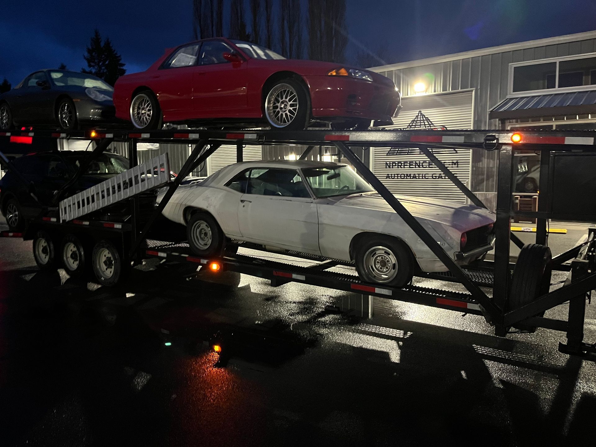 Car carrier truck at night hauling two sports cars, a red car on top and a white car below, on a wet road.