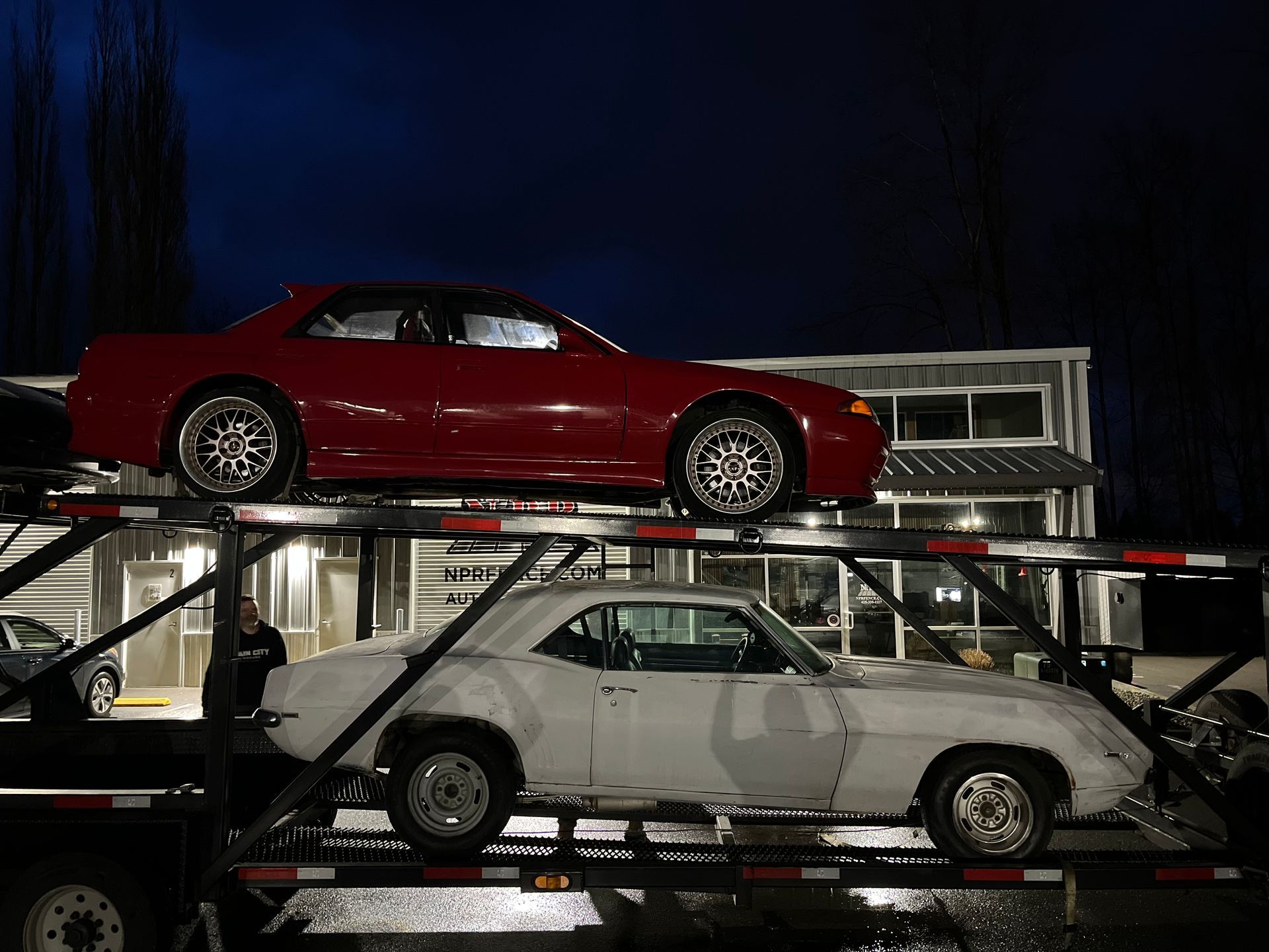 Two classic cars stacked on a transport trailer at night, red coupe above white coupe