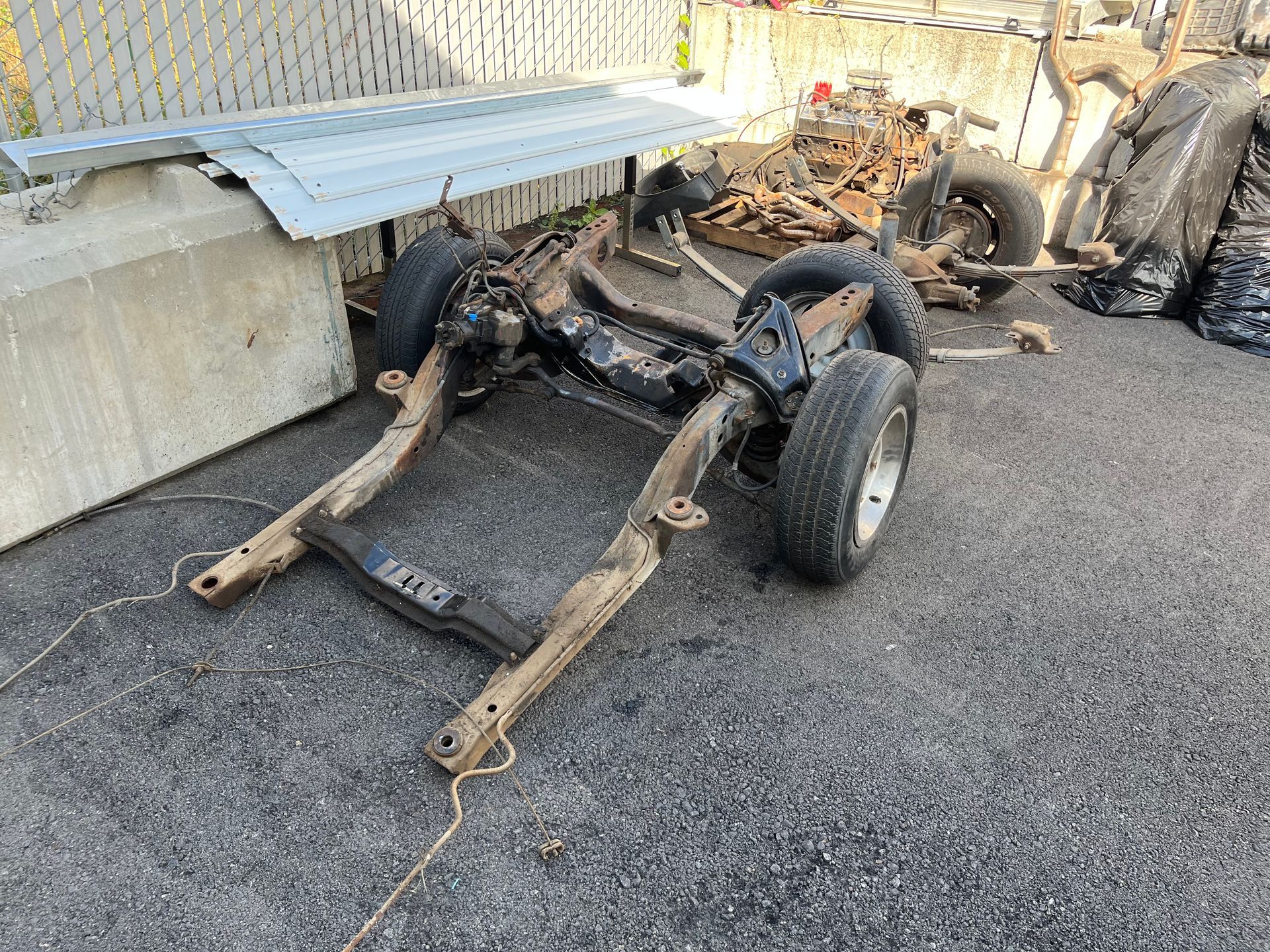 Damaged car chassis and axles on gravel beside a concrete wall and chain-link fence.