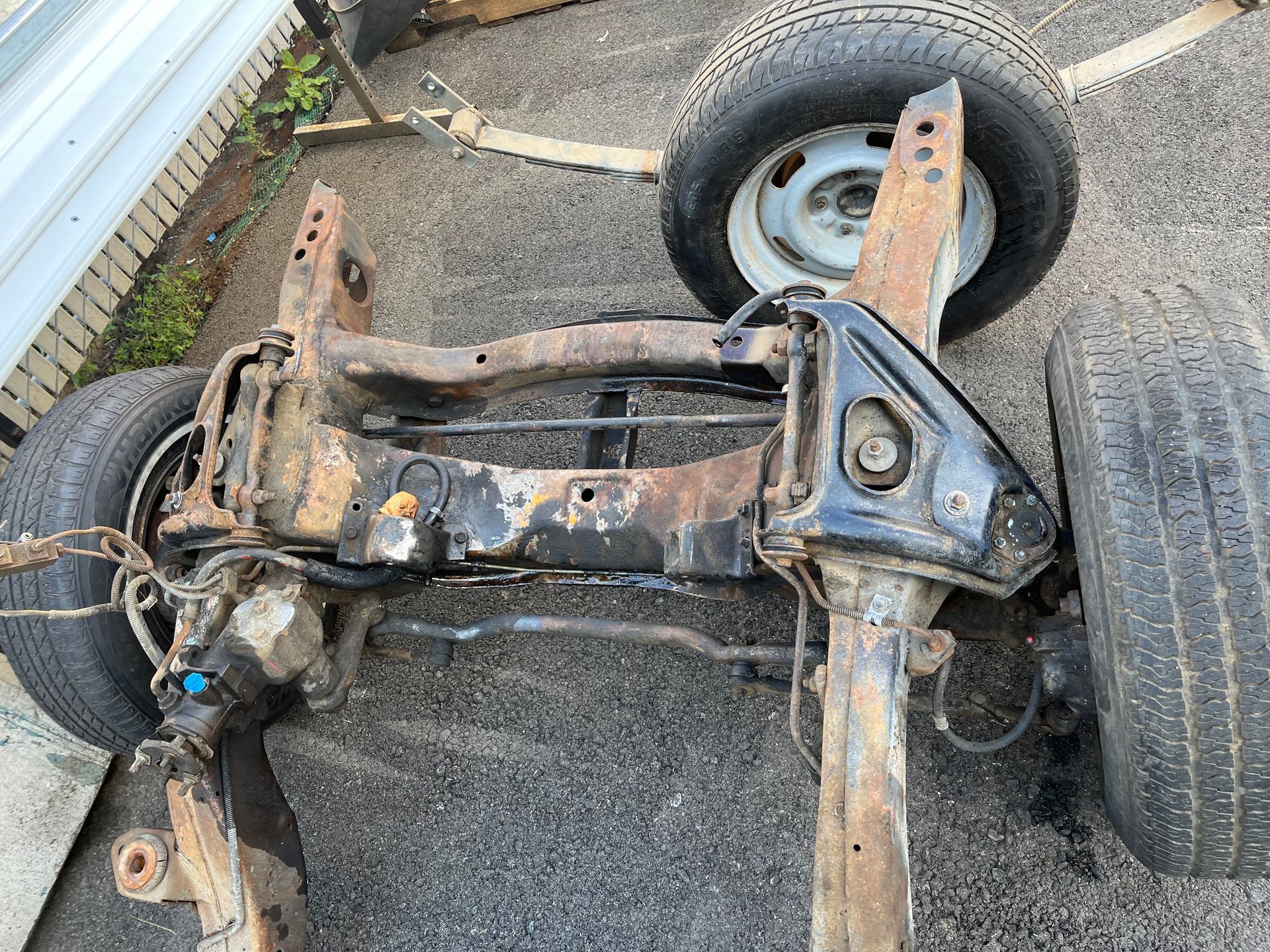 Rusty vehicle chassis and suspension frame with wheels, lying on pavement beside a tire and fence.
