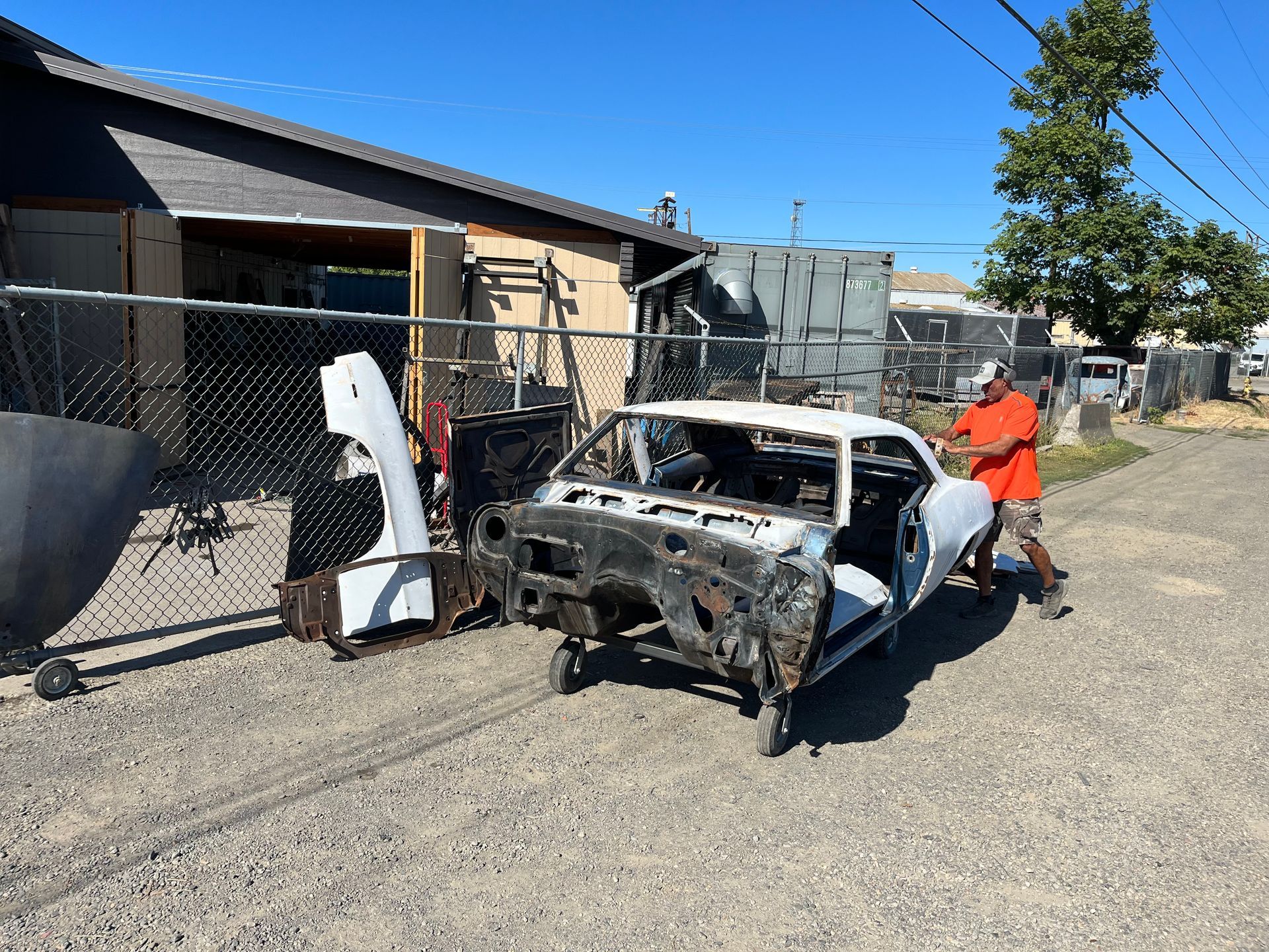 Burned-out car shell on a dirt road, with a person in an orange jacket inspecting it in a residential area