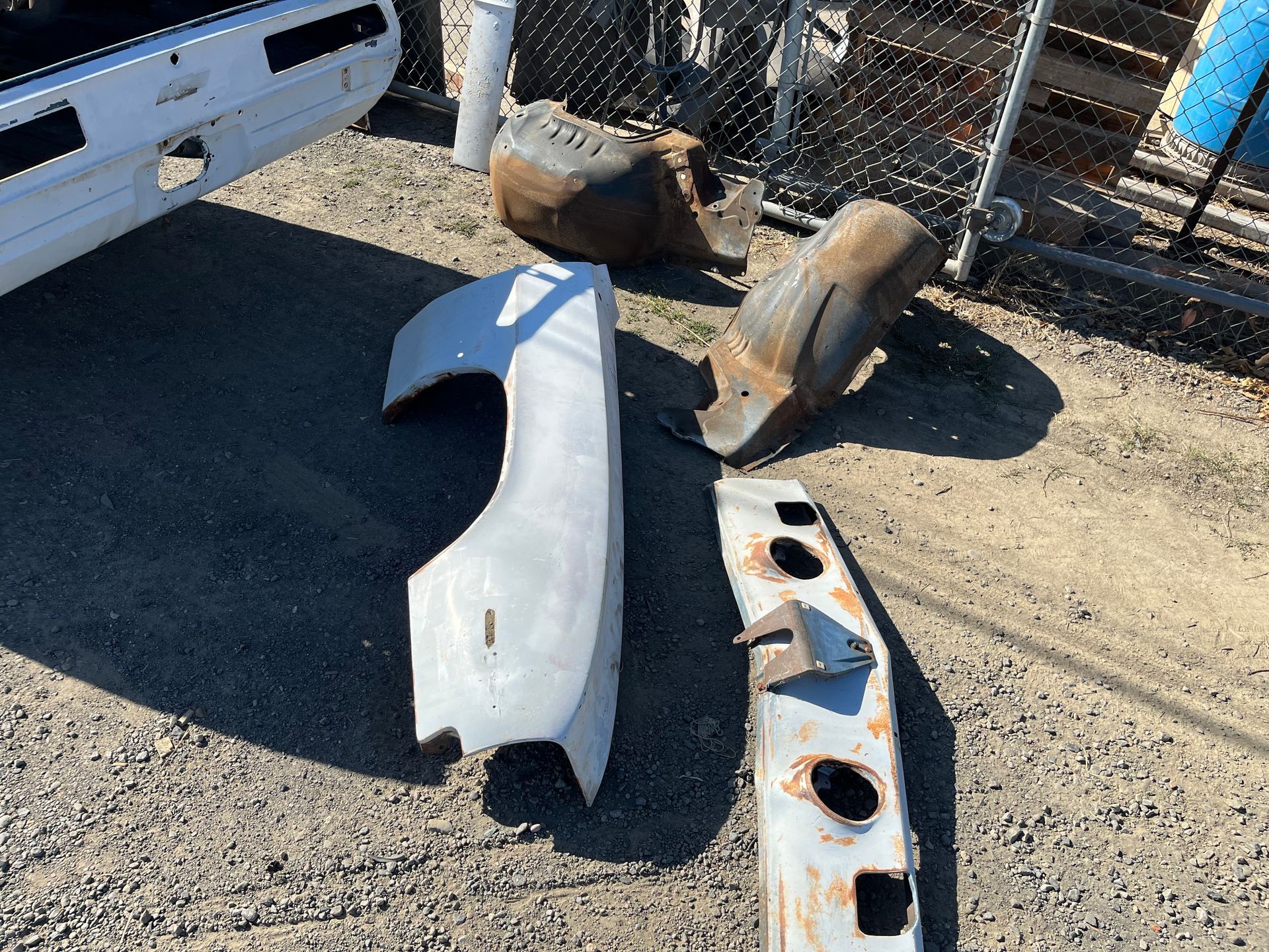 Car bumpers and body panels lying on gravel beside a white vehicle and chain-link fence